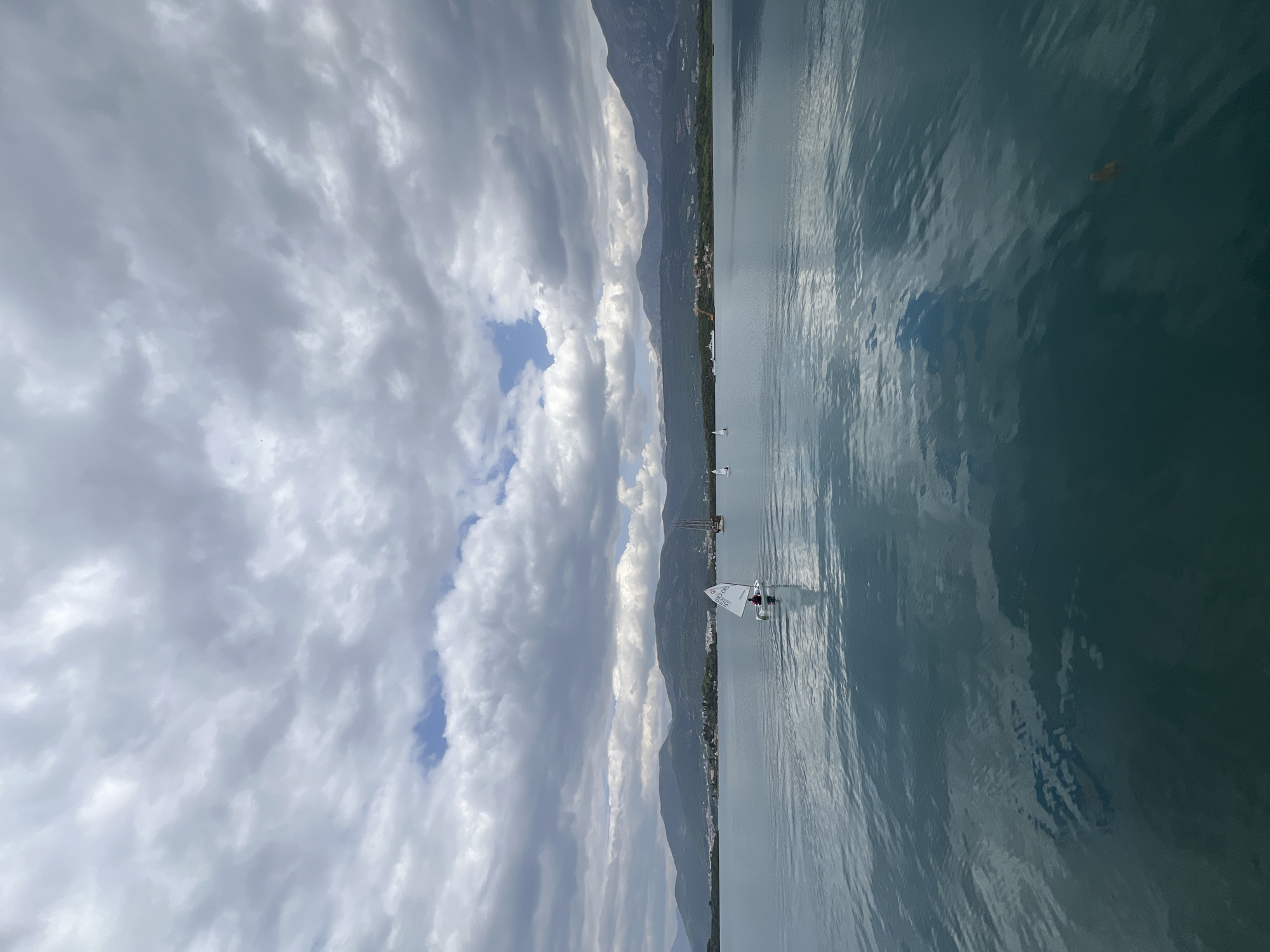 This media file captures a serene waterscape in Đuraševići, Montenegro. The scene is dominated by a calm body of water, likely the Bay of Kotor, with a backdrop of lush green mountains under a mostly cloudy sky.
The primary subject is a small, white sailboat, appearing to be an Optimist dinghy, positioned centrally on the water. A single person, dressed in dark clothing and a red life vest or jacket, is visible inside the boat, engaged in sailing. The sail prominently displays the text "QSC" at the top, a circular logo, and "OPTIMIST" written below it. Further in the distance, to the left of the main sailboat, another smaller white sailboat can be seen.
The water surface is gently rippled and reflects the cloudy sky, creating a soft, diffused light effect. The long stretch of mountains lining the horizon shows signs of human settlement with scattered buildings along their base. The sky is filled with large, grey and white cumulus clouds, though a patch of clearer blue sky is visible towards the left, hinting at shifting weather conditions.
The activity taking place is recreational sailing. The time of day appears to be daytime, possibly late morning or early afternoon, given the ambient light. The weather is calm and partly cloudy. The overall impression is one of peaceful outdoor activity in a scenic natural environment.