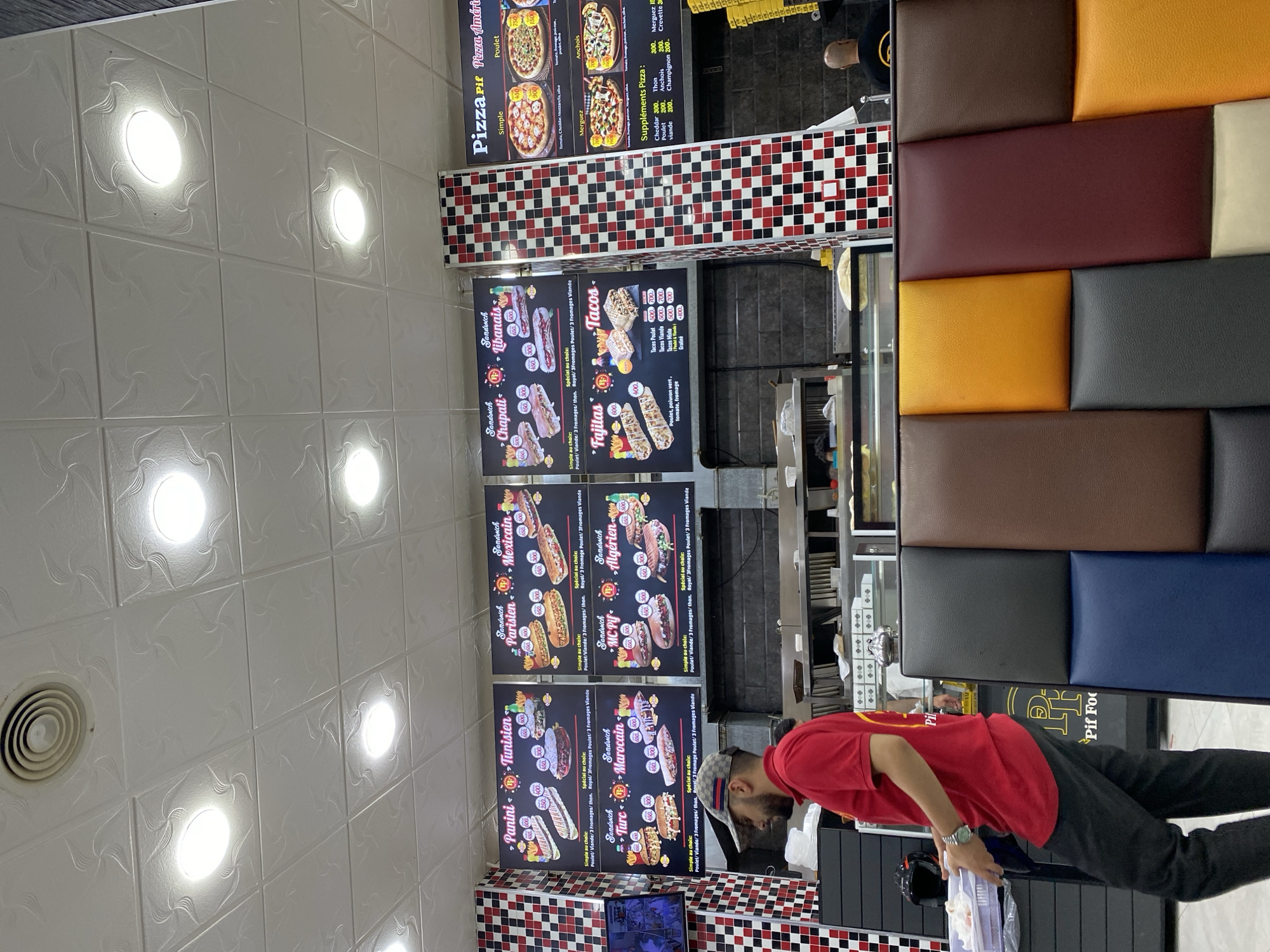 The image captures the interior of a fast-food establishment named "Pif Food" located in Baraki, Algeria. The scene is dominated by a bright, fluorescent-lit ceiling with square tiles. On the walls, there are large menu boards displaying various food items like paninis, sandwiches, fajitas, tacos, and pizzas, each with prices listed in Algerian Dinars. The menus feature images of the food and descriptive text in French and Arabic.
In the foreground, a man wearing a red polo shirt and a patterned baseball cap is seen from the side, carrying a tray with food items. He appears to be a staff member, as suggested by his attire and action of transporting food. Behind him, a counter area is visible with food preparation equipment and displays. To the right, a row of colorful padded seating is arranged, consisting of brown, dark gray, blue, burgundy, orange, and beige panels. In the background, other customers and staff are partially visible. The overall environment suggests a busy, casual dining setting. The decor includes checkered patterns in red, black, and white on some of the vertical surfaces.