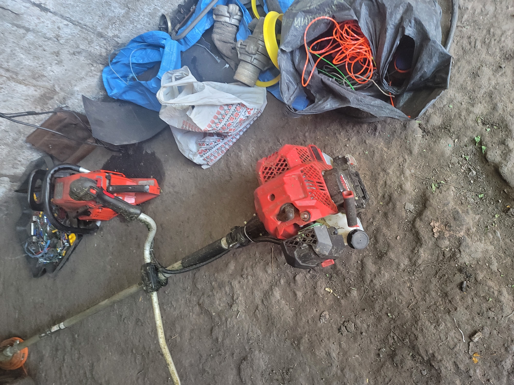 The image depicts a cluttered storage area, likely a shed or garage, in Vovkivka, Ukraine. The ground is covered in dirt and debris. In the foreground, a red string trimmer with a curved shaft and a brush cutter head is lying on the ground, partially obscured by a black plastic bag containing coiled orange and green trimmer line. To the left of the string trimmer, a portion of another string trimmer, with an orange cutting head, is visible. Behind the string trimmer, a red chainsaw is positioned on a black tray filled with various electronic components and wires. To the right of the chainsaw, a brown piece of wood leans against a rough, concrete wall. Further to the right, a blue tarp is draped over some objects, and a white bag with a red and white patterned design is visible. Part of a grey metal pipe or hose is also present near the blue tarp. The overall scene suggests a collection of stored tools and miscellaneous items in a somewhat unorganized setting. There are no people visible in the image.