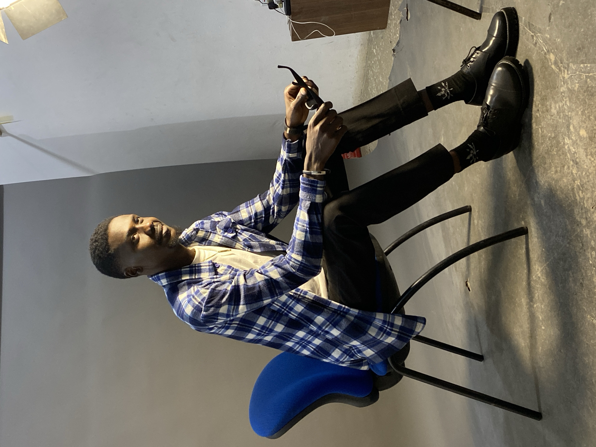 The image depicts a man sitting on a blue office chair in what appears to be an indoor studio setting. He is wearing black trousers, black shoes with patterned socks, a white t-shirt, and a blue and white checkered shirt. He is holding a pair of sunglasses in his hands. The background includes a gray wall and floor, a wooden cabinet, and what seems to be a lighting fixture. There is no definitive visual information to identify the location as specifically being in Jos, Nigeria. The description is based solely on visual information and does not include subjective interpretations or assumptions about cultural context or location.