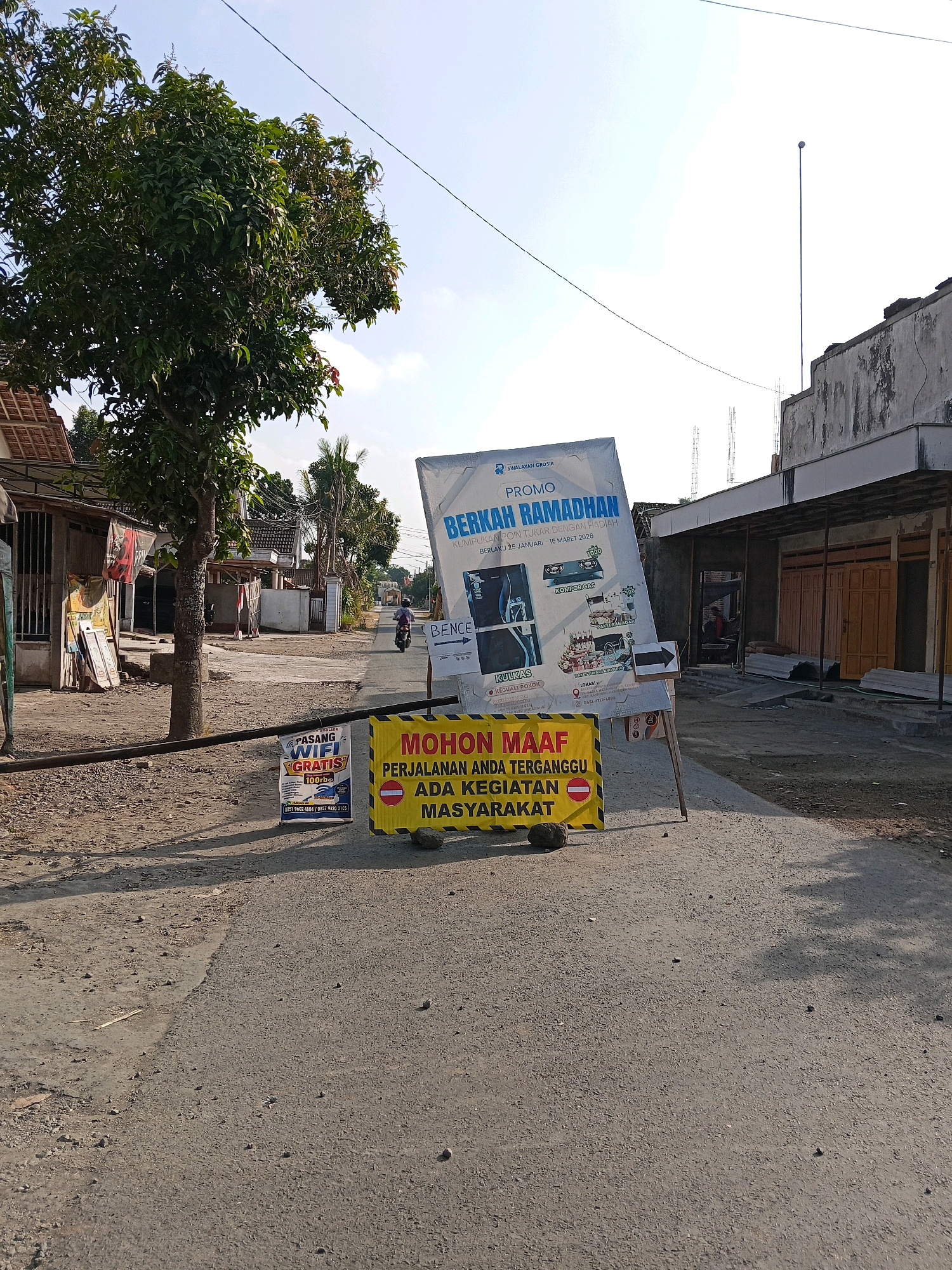 The image shows a street scene in Bence, Indonesia, on a sunny day. In the foreground, a yellow and black striped sign reads "MOHON MAAF PERJALANAN ANDA TERGANGGU ADA KEGIATAN MASYARAKAT," which translates to "Sorry for the inconvenience, there is a community activity." A rock is placed on the sign to keep it from blowing over. To the left, a smaller blue and white sign advertises "PASANG WIFI GRATIS" (Install Free Wifi) with a price of 100,000 Rupiah.
Further down the road, a large promotional banner for "BERKAH RAMADHAN" (Ramadan Blessings) is displayed, featuring various products and a message about exchanging points for gifts. A motorcycle with a rider is visible in the middle ground, moving away from the camera down the street. The street is paved and appears to be a village road. On either side of the street, there are houses and small shops, some of which appear to be under construction. A large tree with lush green foliage stands to the left of the road, casting a shadow. The sky is clear with a few wispy clouds. The overall impression is a quiet, semi-urban street in Indonesia during the day.