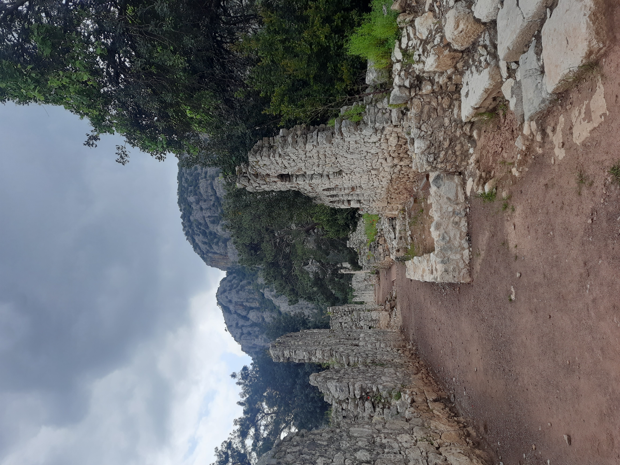 The image displays an ancient archaeological site, likely located in Kumluca, Turkey. The scene is dominated by the weathered remnants of stone structures, specifically, thick stone walls constructed from irregular, light-colored blocks. These ruins run along the right side of a reddish-brown dirt path, which appears to be a well-trodden route through the site. More ruins and dense green foliage are visible to the left of the path, with large trees overarching parts of the scene from the upper left.
In the background, imposing, rugged mountains rise under an overcast sky filled with heavy grey clouds, indicating a cloudy daytime. These mountains are partly covered with green trees and vegetation, blending with the dense greenery that also surrounds the ruins in the midground. No individuals or specific activities are depicted, giving the scene a serene and contemplative atmosphere. The image emphasizes the historical antiquity of the ruins and their integration with the natural environment. No visible text is present.