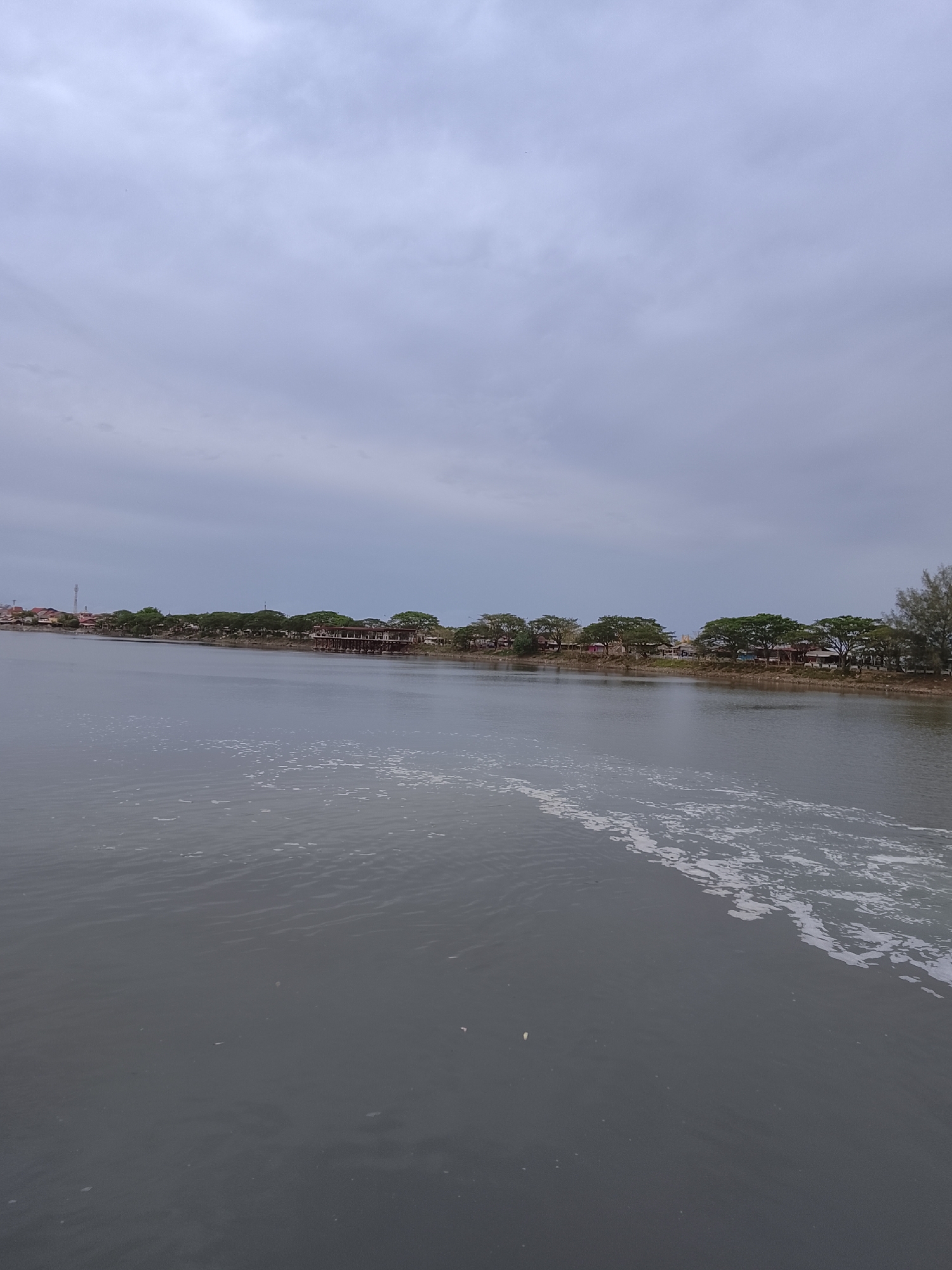 The image depicts a body of water, likely a river or bay, with a visible shoreline across the water. The water's surface is mostly calm with some ripples and a line of foam or wake extending from the foreground toward the middle distance. The shoreline is lined with trees and some buildings, possibly houses or commercial structures. The sky is overcast and grey, suggesting a cloudy day. The image is taken in Lhokseumawe, Indonesia. There are no people visible in the shot. The primary subjects are the water, the shoreline, and the sky.
