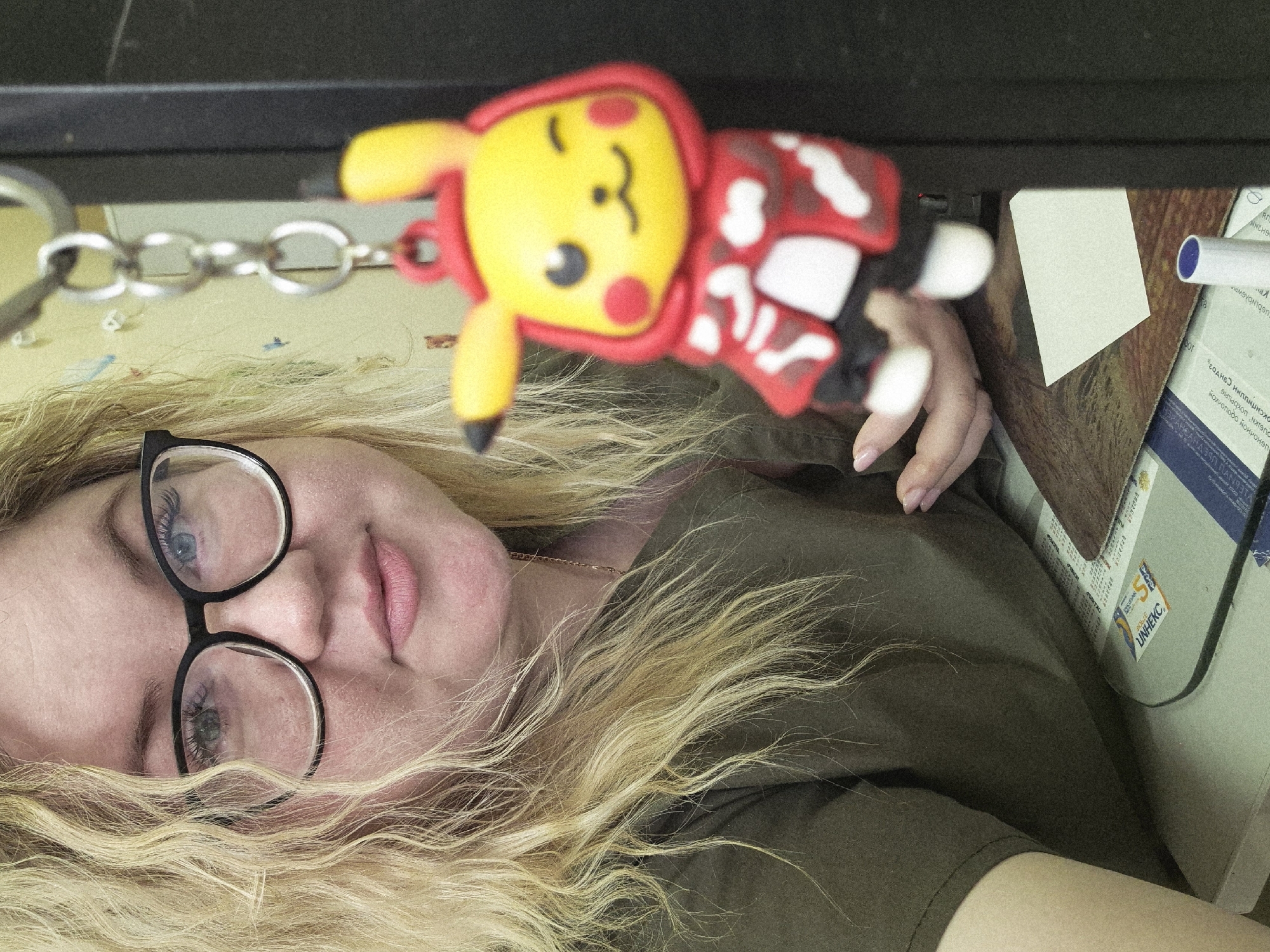 A woman with long, curly blonde hair and black-rimmed glasses is lying down. She is wearing a dark green shirt. Above her is a Pikachu keychain with a red and white patterned covering. The keychain is attached to a silver chain. Behind the keychain, there is a dark surface and a desk with papers and a blue pen. The location is not identifiable as being in Фоки, Russia.