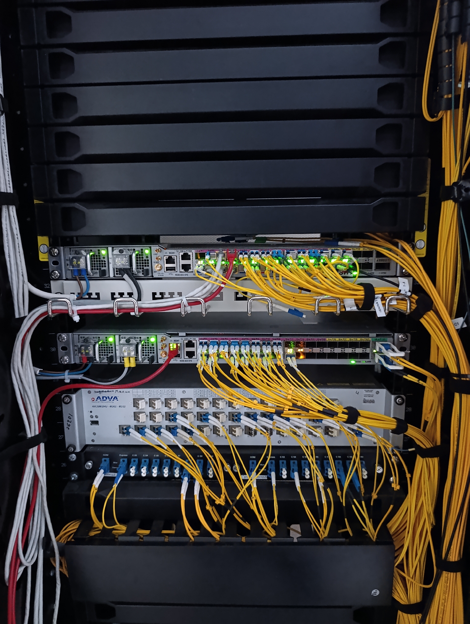 This is a close-up, eye-level, indoor, static, full shot of a server rack filled with networking equipment. The server rack is predominantly black with several shelves holding various devices. Multiple yellow fiber optic cables are prominently routed through the rack, bundled with black zip ties. Some of the devices have green indicator lights on. There are also white and red cables connected to some of the equipment. The background shows the dark interior of the rack. The image does not contain any people or discernible actions. The scene is clean and organized, typical of a data center or telecommunications equipment room. The location in the city of Dubăsari town council, Moldova is not visually evident from the image itself.