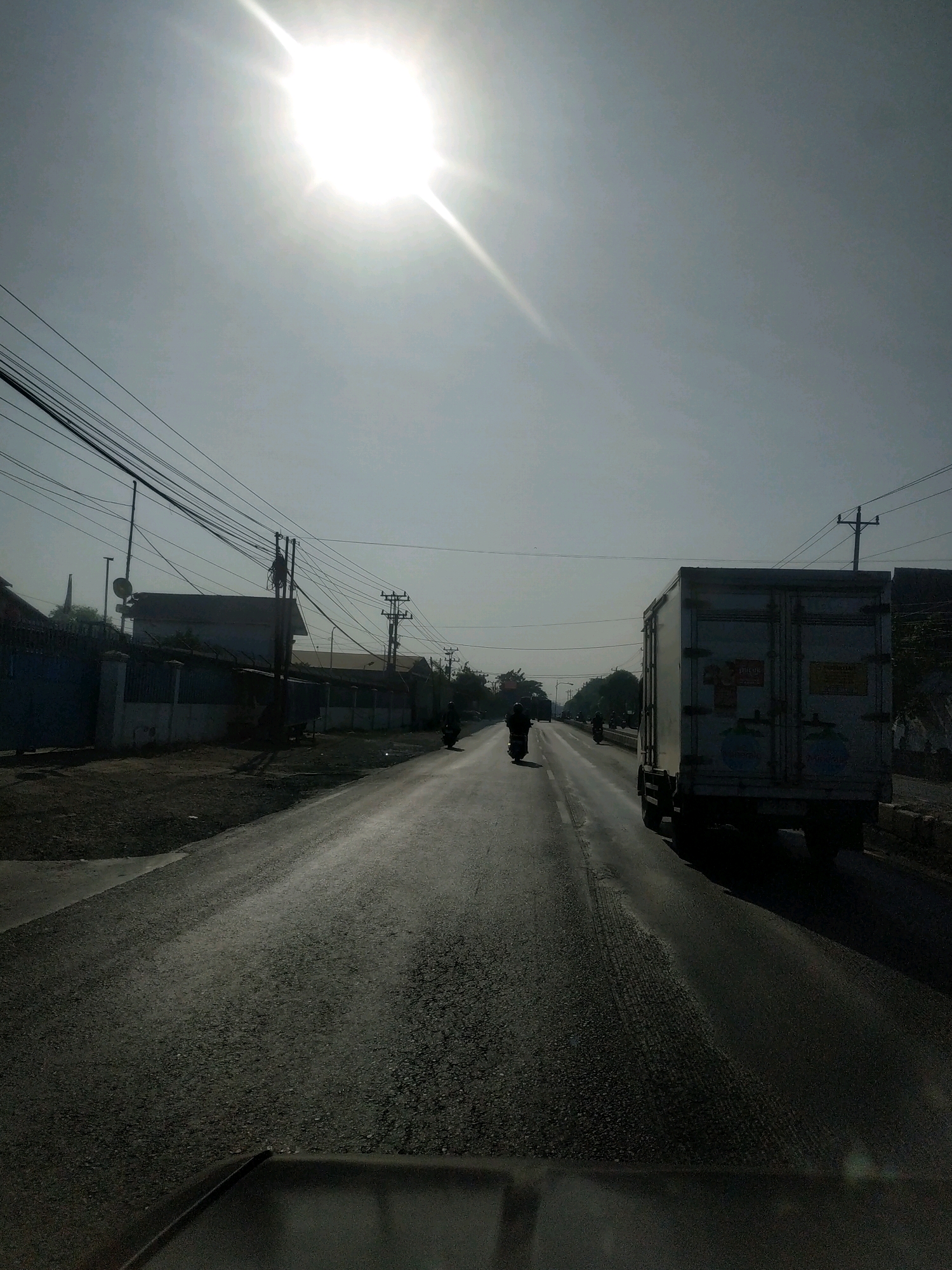 The image depicts a sun-drenched road in Padaharja, Indonesia. The bright sun is high in the sky, casting a strong glare. On the right side of the road, a white box truck is driving away from the camera. Further down the road, several motorcycles are visible, also moving away. The left side of the road features buildings and a fence with barbed wire. A dense network of utility lines stretches across the sky, supported by poles. The road surface is asphalt, with some patches of wear and tear. The scene suggests a typical day on a road in an urban or semi-urban area of Indonesia.