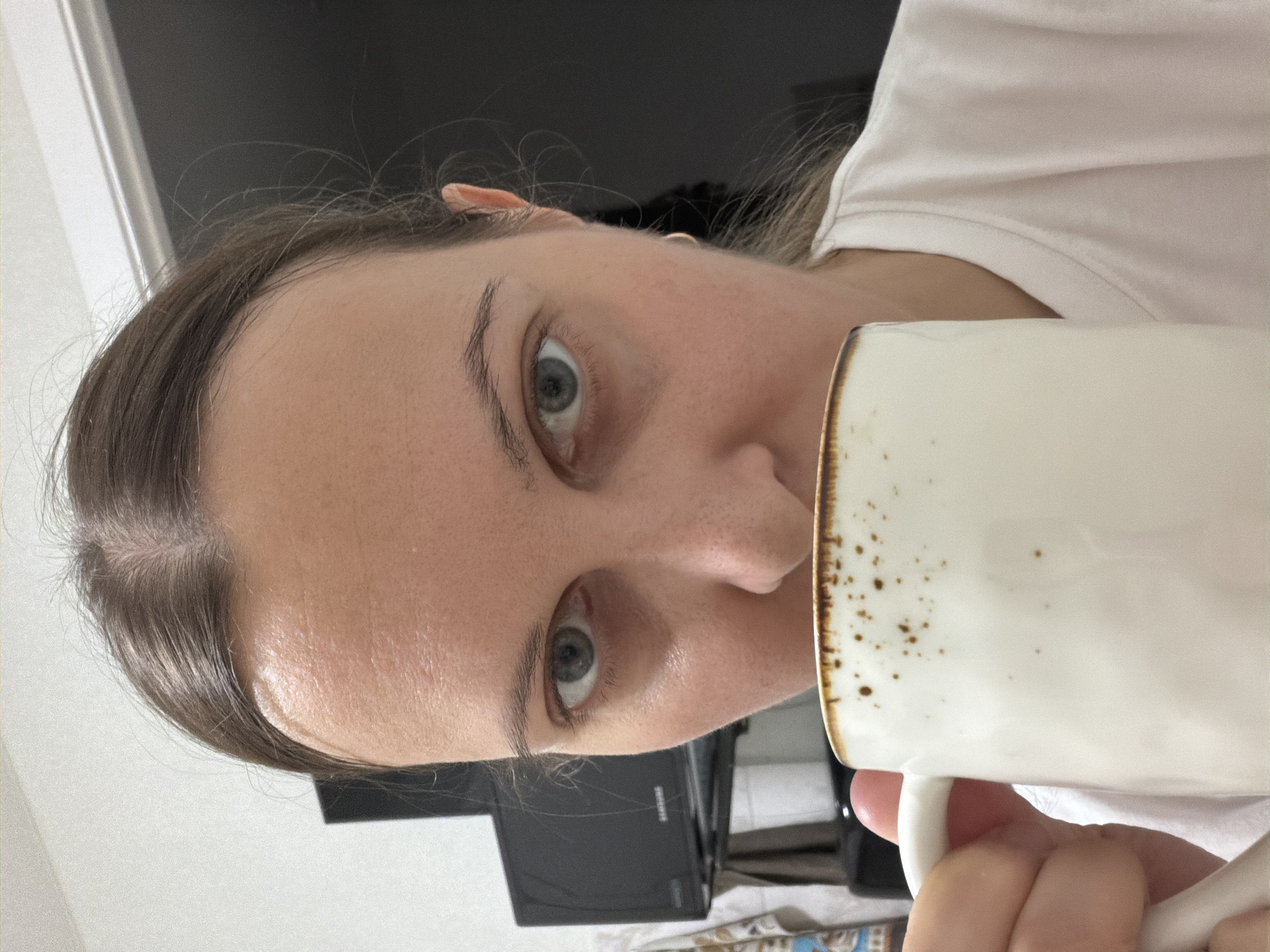 A close-up, head-and-shoulders shot captures a young Caucasian woman holding a white mug with a speckled brown rim to her lips, mid-sip. Her dark brown hair is pulled back, revealing a fair complexion with visible pores and a few freckles. Her light gray eyes are directed towards the camera with a neutral expression. A dark, flat-screen television is visible in the background, along with the corner of a white wall. The lighting is soft, suggesting an indoor setting, possibly a home. The woman is wearing a light pink t-shirt. The image is cropped such that the lower portion of her face and the top of the mug are prominent. There is no discernible information about the specific location in Izhevsk, Russia, beyond it being an indoor residential or personal space.