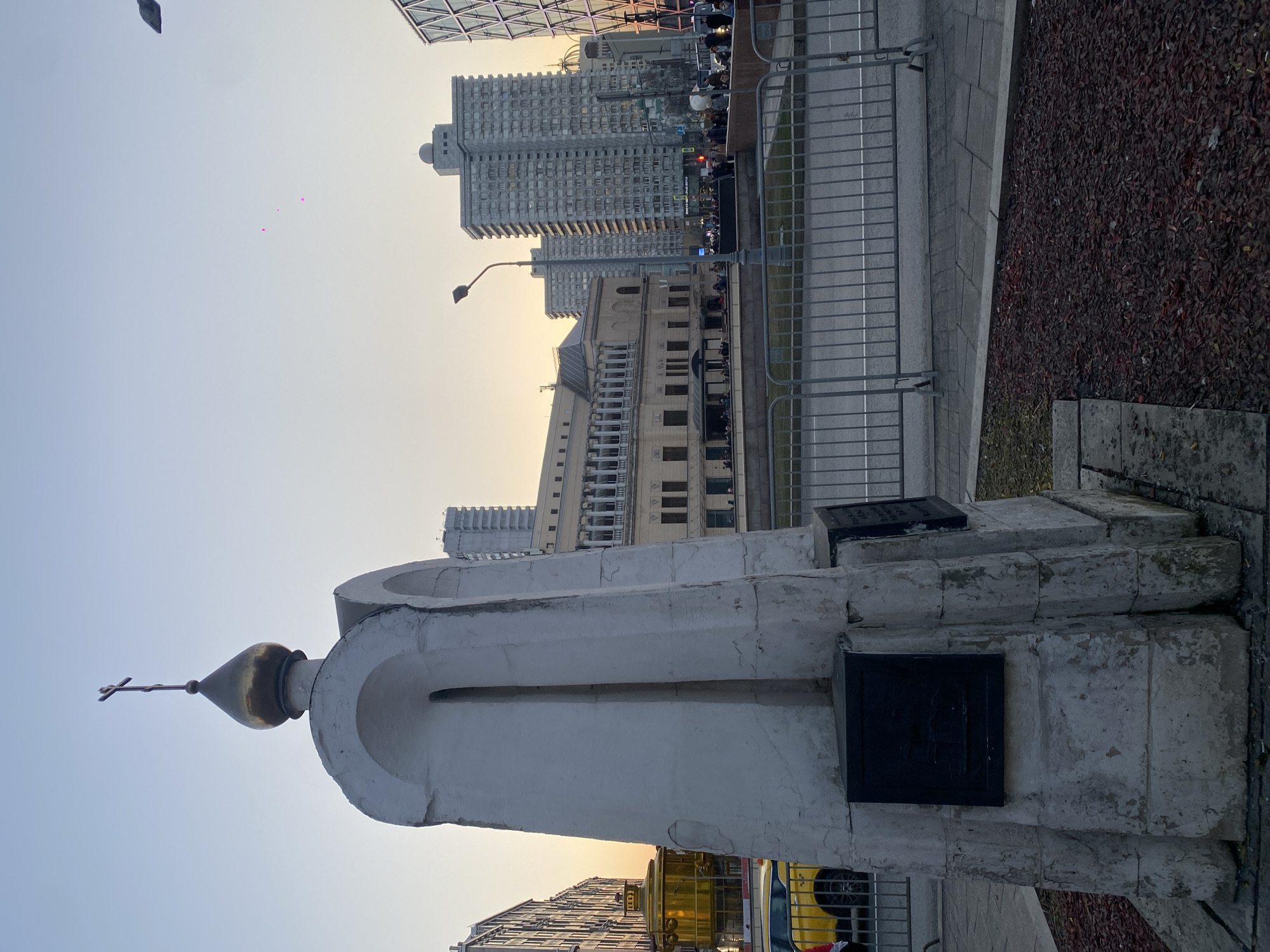 The image depicts an urban street scene in Moscow, Russia, specifically on Bolshaya Yakimanka Street. In the foreground, a weathered white stone monument with a golden onion dome and an Orthodox cross at its apex stands on a bed of reddish-brown wood chips. Two dark, rectangular plaques are affixed to its base.
Behind the monument, grey metal crowd control barriers run horizontally, separating the foreground from a multi-lane asphalt roadway with white markings. A black streetlamp is positioned further back, between the road and a distant embankment.
In the midground, beyond the roadway, a large, cream-colored building with multiple stories, numerous windows, and a prominent upper section featuring balconies and arched openings is visible. Below this building, on what appears to be a promenade or embankment, several individuals are present, some congregated in groups.
The background features several tall, grey, multi-story buildings, characteristic of modern urban architecture. To the far right, part of a geometrically patterned, glass-fronted building is visible. To the far left, a yellow taxi is partially obscured by the monument, next to another building.
The sky exhibits a gradient, transitioning from a soft orange-yellow near the horizon to a lighter blue overhead, indicating either sunrise or sunset. Three small, faint pink specks are visible in the upper portion of the sky.