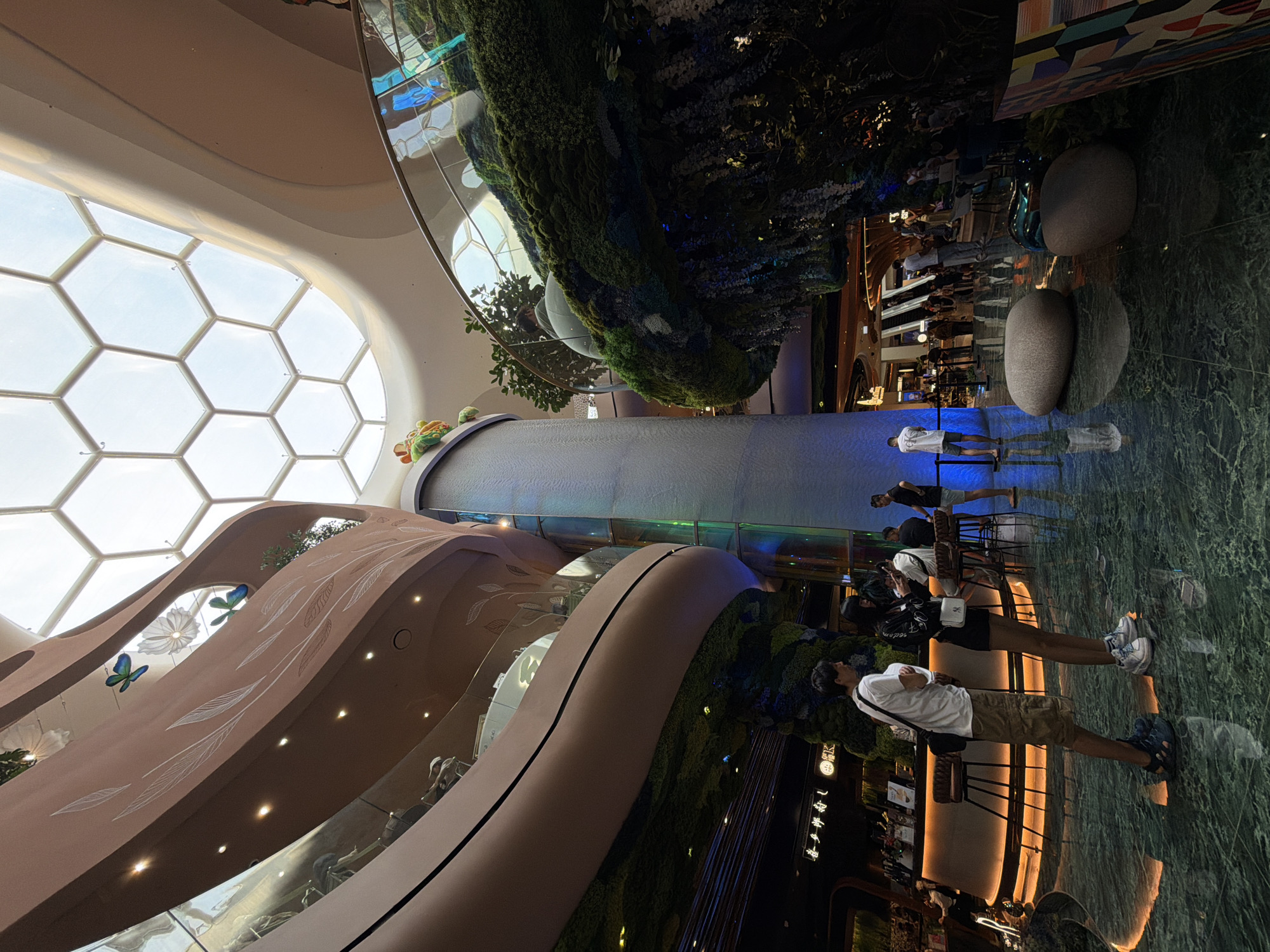 The image depicts the interior of a modern shopping mall or building, possibly in Bangkok, Thailand, suggested by the architecture and ambiance. The space features a large, open atrium with a honeycomb-patterned glass ceiling allowing natural light to enter. A prominent vertical water feature or cascade is visible, surrounded by lush greenery and plants, creating a garden-like atmosphere.
Several people are present in the scene. Some are standing and appear to be observing the water feature or the environment, while others are sitting at tables, likely in a restaurant or dining area. They are dressed in casual clothing.
The environment is characterized by curved, organic architectural elements, possibly escalators or walkways, decorated with nature-inspired designs like leaves and butterflies. The flooring appears to be polished marble or stone. The lighting is a mix of natural daylight and artificial illumination from overhead fixtures. The overall impression is one of a high-end, modern, and nature-integrated space.
