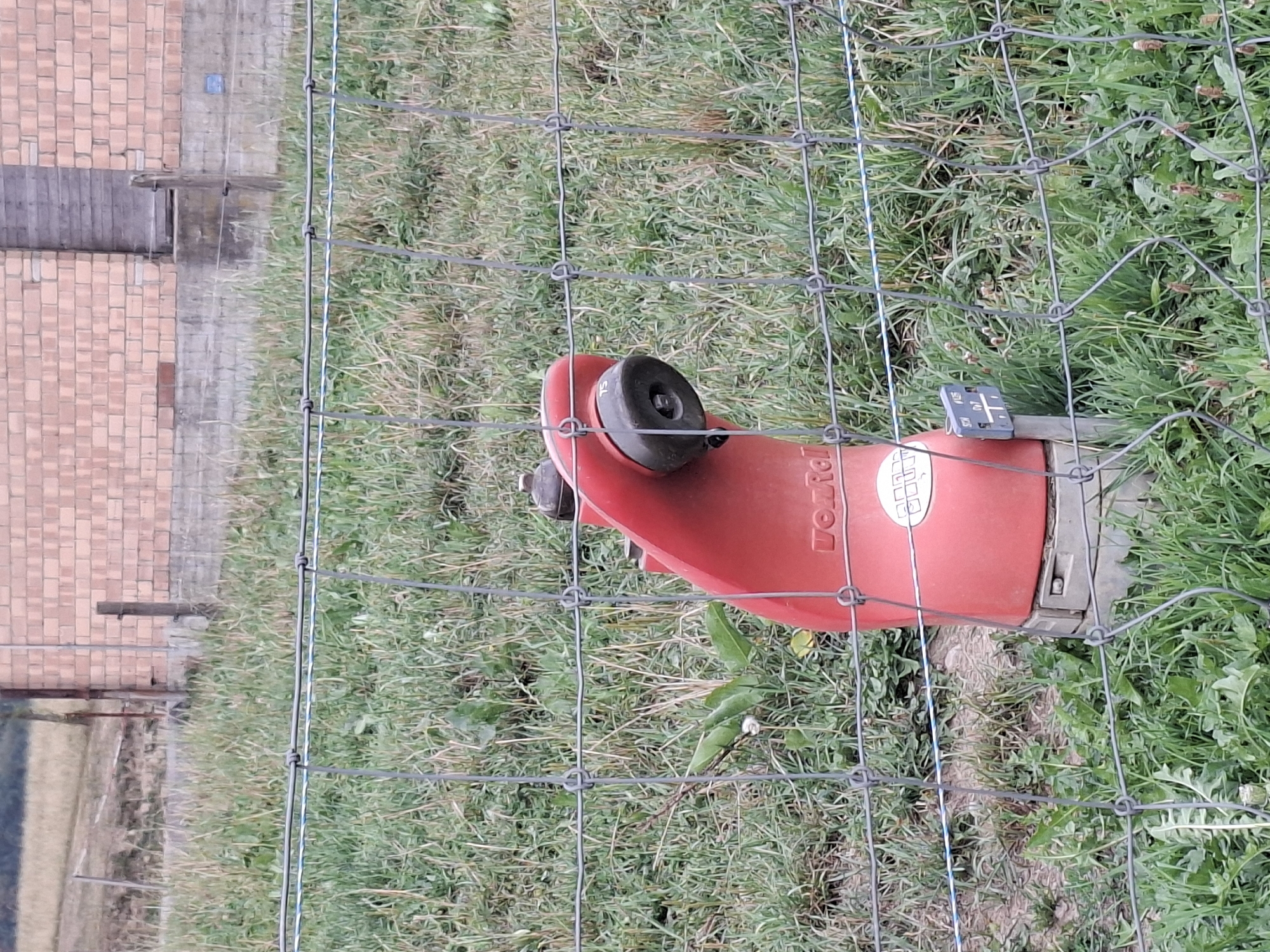 A red fire hydrant stands behind a wire fence in a grassy field. The hydrant is red and has a black nozzle. There is a small blue sign attached to the hydrant. In the background, there is a brick building. The field is covered in green grass and some brown dirt. The sky is not visible in the image.