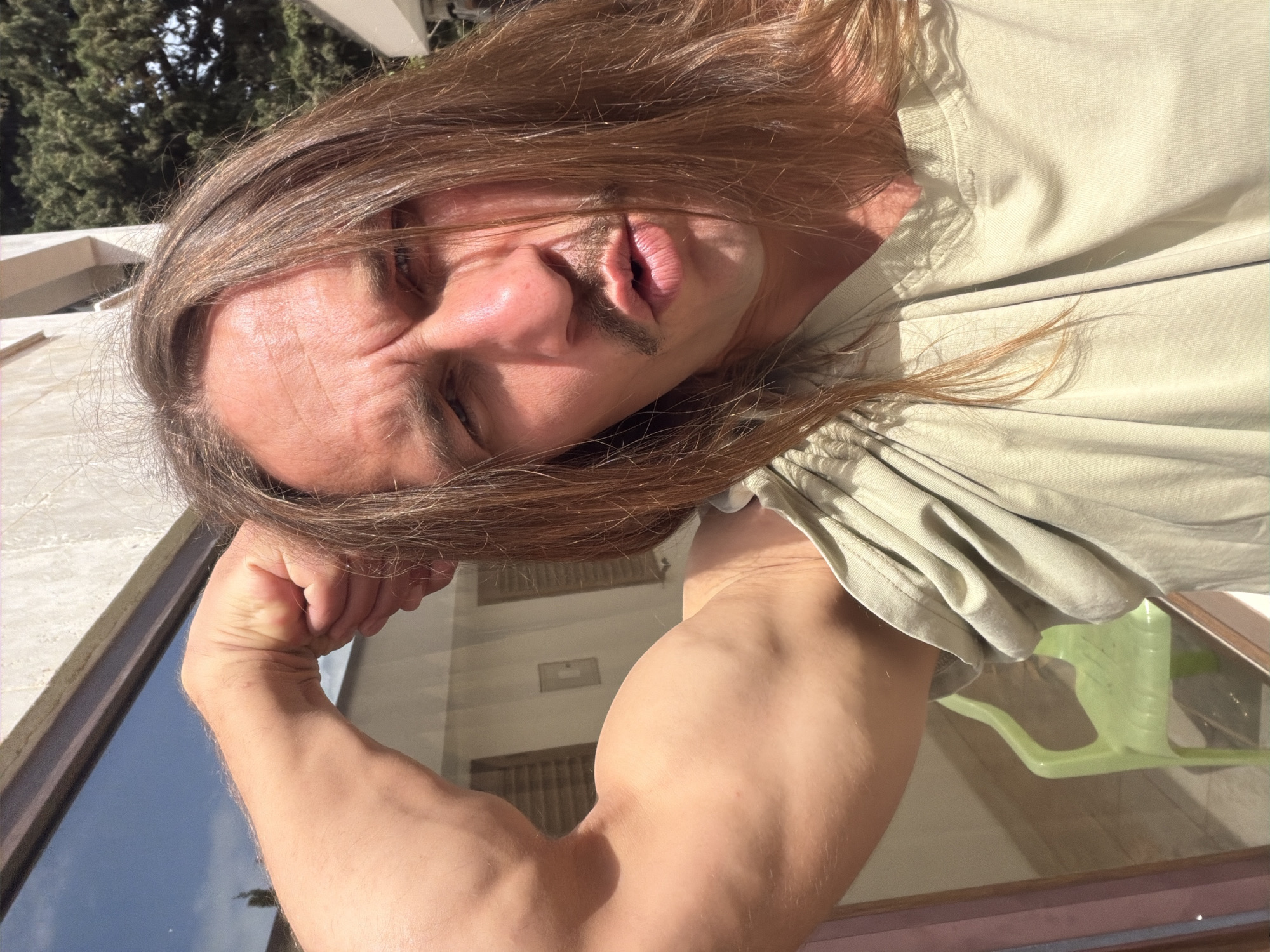 A man with long brown hair and a mustache is flexing his right bicep, which is prominently displayed in the foreground. He is wearing a light-colored t-shirt. The scene appears to be outdoors on a sunny day, with a tree visible in the upper left corner. The reflection in the window behind him shows buildings. A green chair is partially visible in the bottom right corner. The location is Petrovac centar, Montenegro.