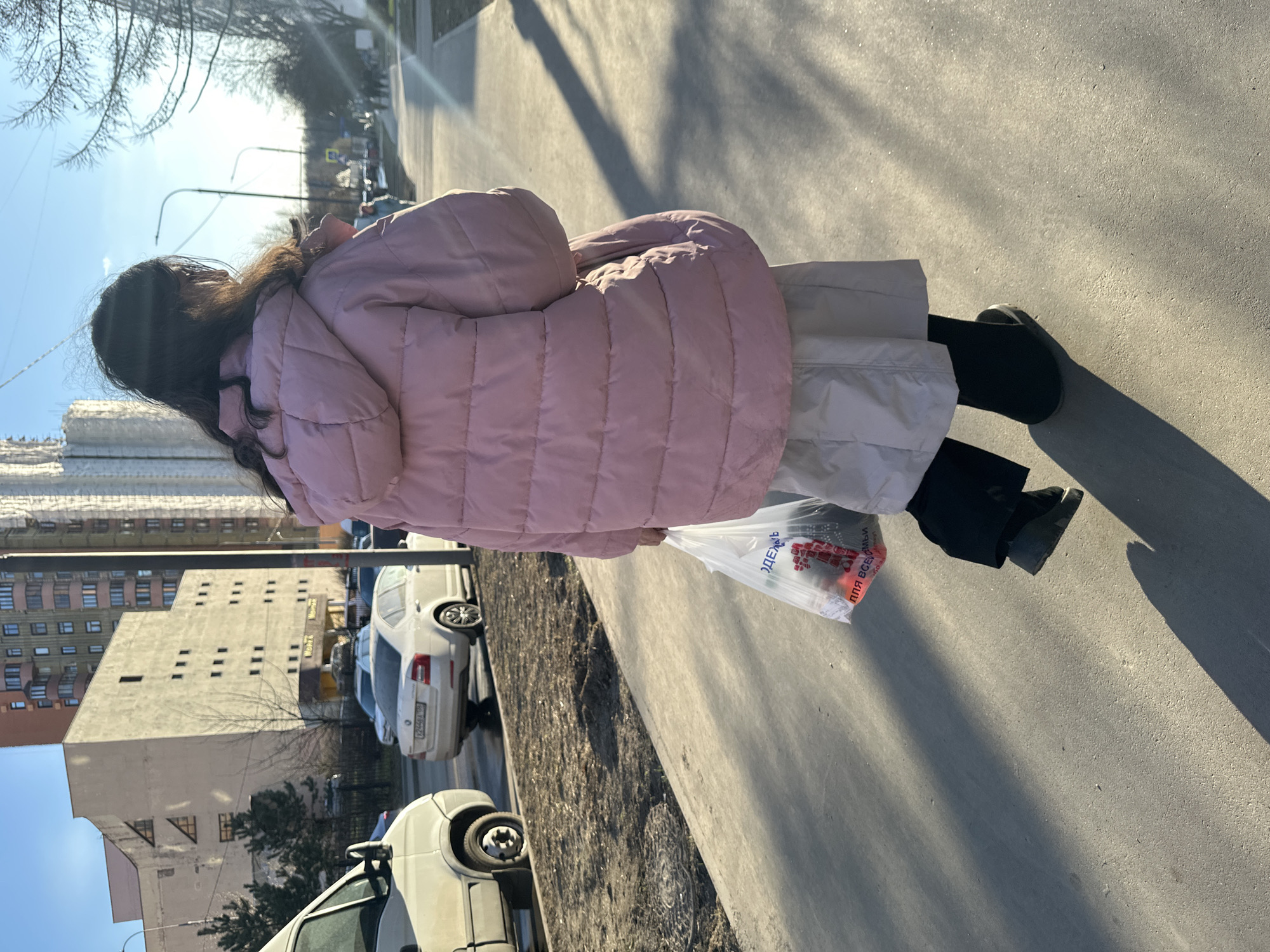 A woman is walking on a sidewalk in Moscow, Russia. She is wearing a pink puffer jacket, a light-colored skirt, and black shoes. She is carrying a plastic bag in her right hand. The buildings in the background are typical of Moscow's urban architecture. Cars are parked along the side of the road. The day appears to be sunny.