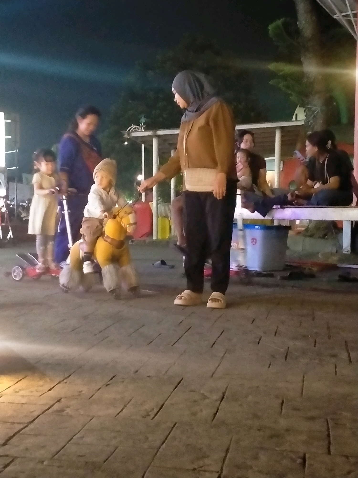 In an outdoor, nighttime setting in Batu, Indonesia, several individuals are present. In the foreground, a child wearing a light-colored outfit and a beige beanie is seated on a yellow toy horse with shaggy gray legs, propelled by a string held by an adult. To the left, another child in a light-colored dress stands on a scooter with three wheels. A woman in a blue garment stands behind the child on the scooter. A woman wearing a brown jacket, black pants, and a headscarf, holding a white sash around her waist, stands to the right of the toy horse, appearing to supervise. In the background, a group of people are seated on a bench. The ground is paved with rectangular stones. Lights are visible in the background, and trees and a structure with a white roof are also present.