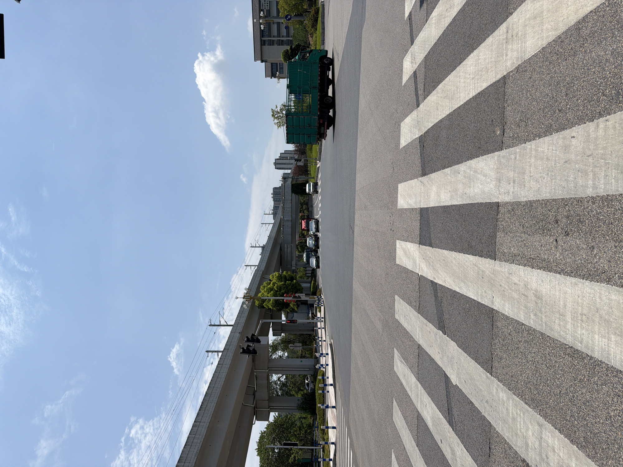 This is a medium shot, eye-level photograph of a busy street intersection in Jiangjin District, Chongqing, China, on a bright, sunny day. The foreground is dominated by a pedestrian crossing with thick white stripes painted on dark asphalt. In the middle ground, multiple vehicles are present. A large, green truck with a caged rear is turning right. To the left, several cars and a white van are proceeding through the intersection. In the background, a modern elevated railway structure with overhead wires traverses the scene. Beyond the railway, buildings and trees line the horizon under a pale blue sky with scattered white clouds. Traffic lights are visible at the intersection, indicating the flow of traffic. The overall impression is an urban environment with active transportation infrastructure.