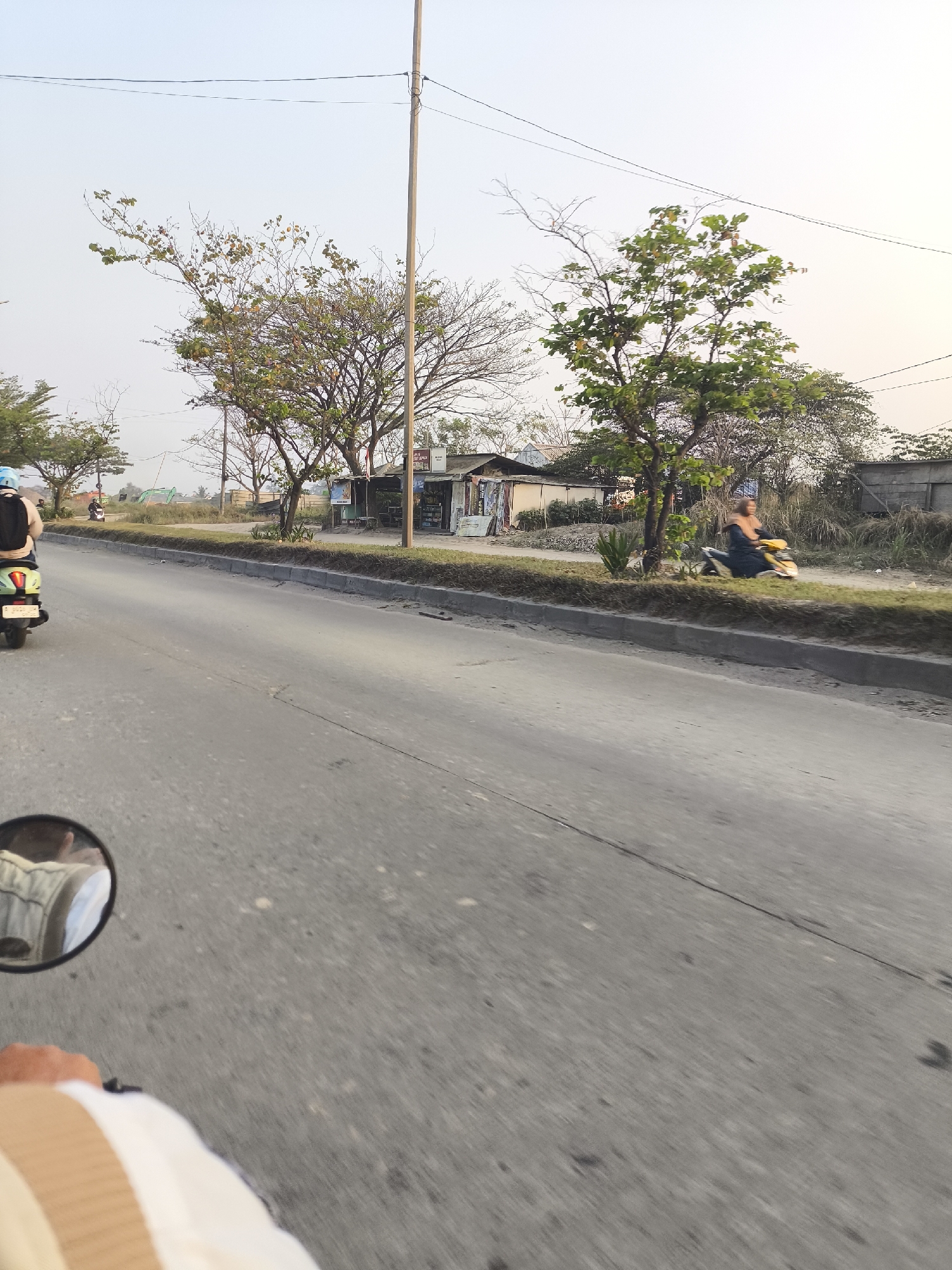 The image captures a scene on a road in Cilegon, Indonesia, likely during the late afternoon given the soft, diffused light. The perspective suggests the photo is taken from the viewpoint of someone riding a motorcycle, as evidenced by the motorcycle's handlebar and side mirror in the foreground. The road surface is asphalt, with a clear dividing line visible.
To the right of the road, a grassy embankment is present, bordered by a concrete curb. Beyond the embankment, there are several structures that appear to be small shops or informal dwellings, constructed from corrugated metal and wood. Trees, some with sparse foliage, line the area behind these structures, contributing to a somewhat rural or semi-urban landscape. In the far background, a fence and some agricultural fields or undeveloped land are visible.
Two individuals are depicted on motorcycles. On the left, a person wearing a blue helmet and a black backpack is riding a light-colored scooter, moving away from the camera. Further down the road, on the right side, another individual is riding a yellow motorcycle, also moving away. The rider appears to be wearing a dark top and is partially obscured by their vehicle and motion blur. Overhead, power lines are strung between poles, a common sight in Indonesian cities. The overall atmosphere is one of everyday life on a roadside in a tropical climate.