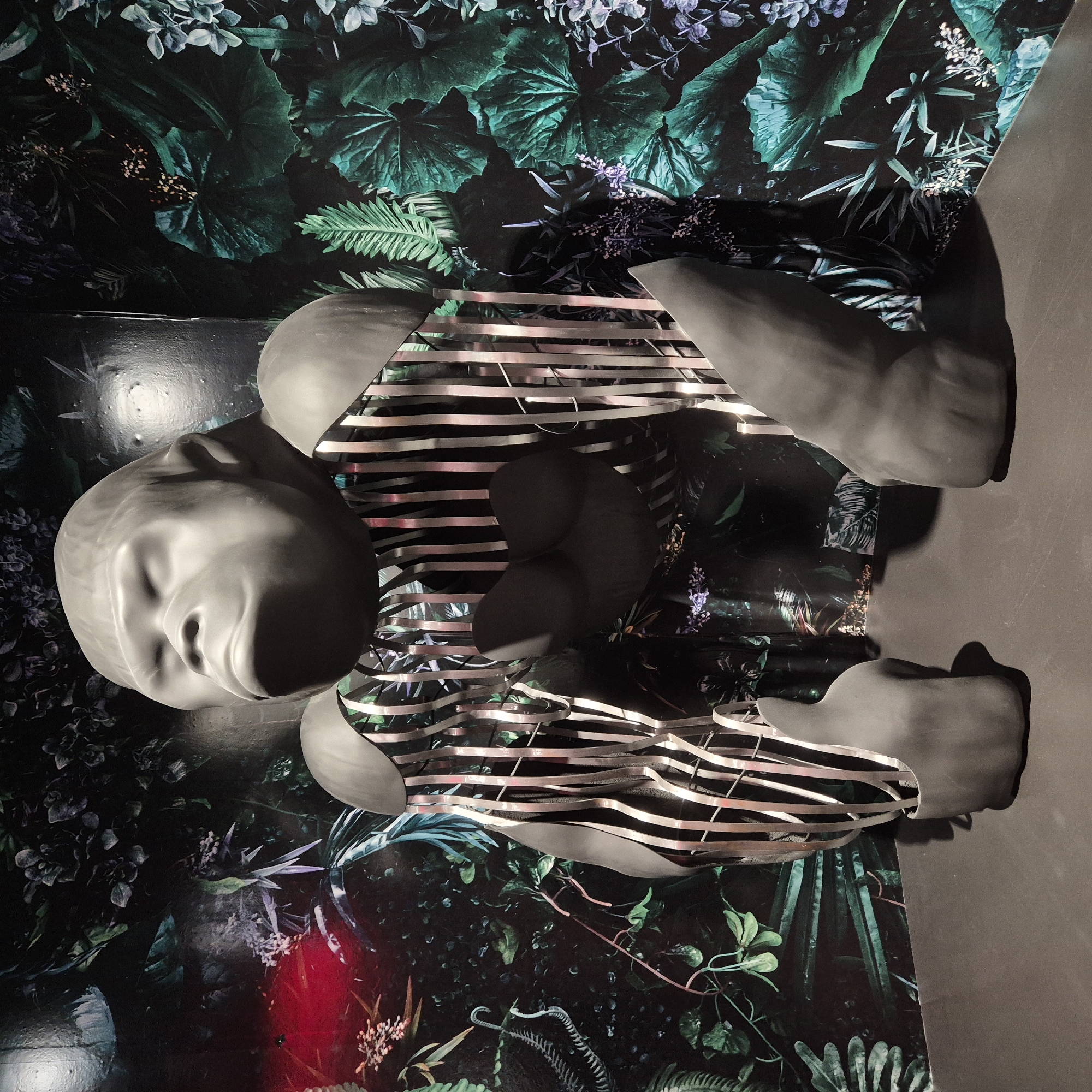 A mannequin lies prone on a backdrop featuring a dark, lush floral pattern. The mannequin's torso is partly encased in a series of horizontal, light-reflective bands, giving it a fragmented appearance. The scene is situated in Strasbourg, France. The background's botanical design includes large leaves, small flowers, and fern-like fronds. The mannequin is gray-toned, with a smooth, featureless face and limbs, suggestive of sleep or death.