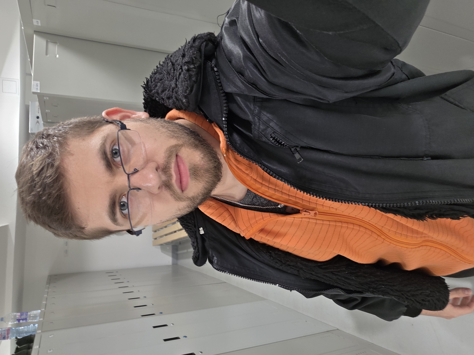 A man stands in a brightly lit, institutional-looking room in Plovdiv, Bulgaria. He is fair-skinned, with brown hair, a beard, and wears glasses. He is dressed in an orange zipped hoodie under a black jacket with a furry collar. The room features rows of metal shelving with slots or perforations running along the sides. Bottles of water are visible on one of the lower shelves. A wooden slatted structure, possibly a bed frame, is also visible in the background.