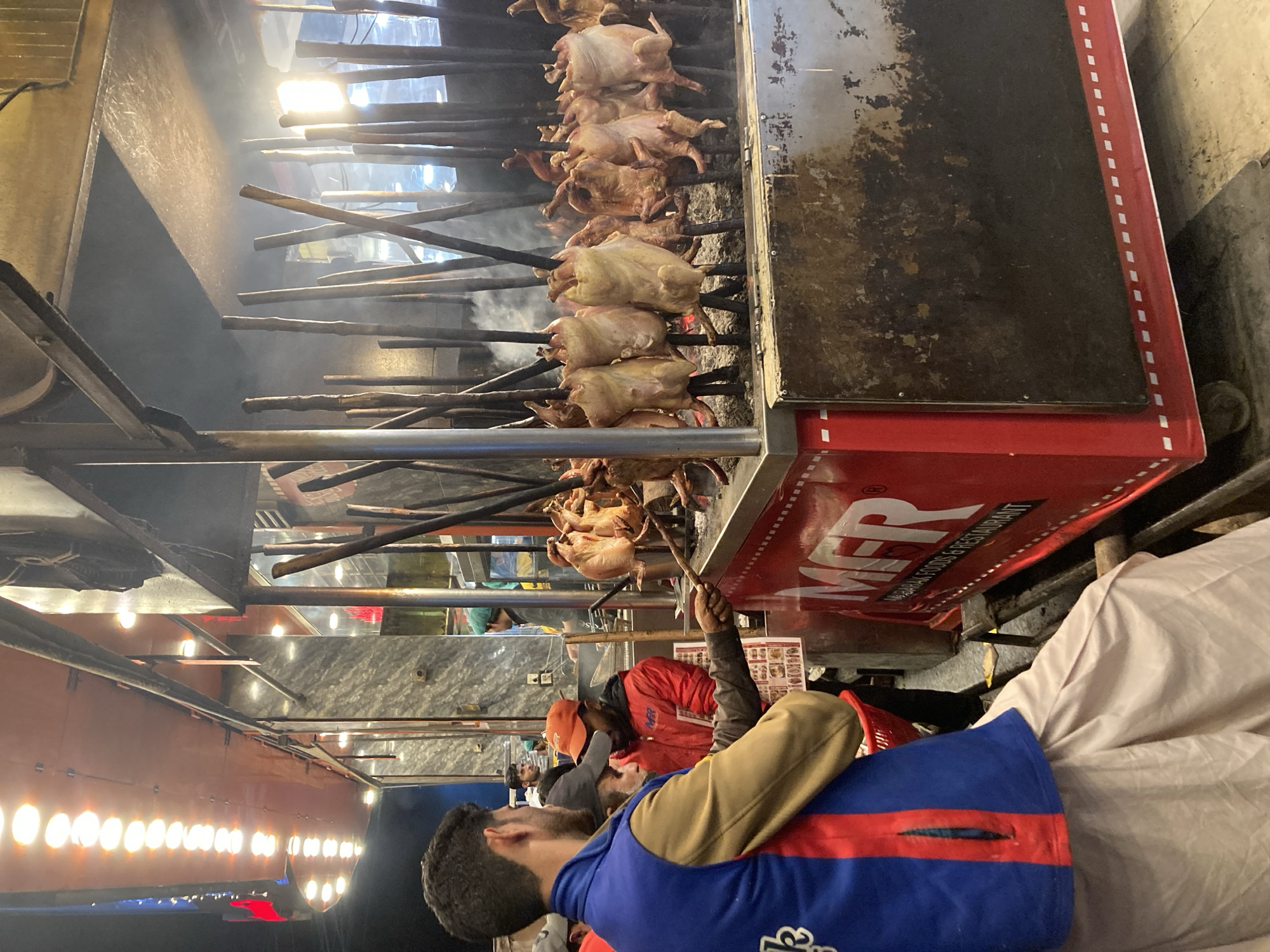 The image depicts an outdoor street food stall specializing in roasted chicken in Quetta City Tehsil, Pakistan, at night. In the foreground, a male individual with dark hair is seen from behind, wearing a blue and red vest over a tan shirt and light-colored shalwar kameez trousers. He extends his right arm toward the stall, holding a long, dark stick. Adjacent to him, another male wears an orange baseball cap and a red jacket displaying the "MFR" logo.
The central subject is a large, red metal food stall, labeled "MFR Mubarak Foods & Restaurant." The stall features an open-air roasting area filled with numerous whole chickens impaled vertically on long wooden or metal skewers. These chickens are cooking over visible embers or an open flame, generating a significant amount of smoke that rises into the air. Some chickens appear raw or partially cooked, while others exhibit a more roasted appearance.
Behind the stall, a permanent building with grey marble-patterned walls and glass panels is visible, illuminated from within. Several individuals are present inside this structure; one person wears a green cap, suggesting staff attire. Overhead, a large awning or canopy features a prominent row of circular, bright artificial lights. An intense light source is also visible directly behind the roasting chickens on the right side of the frame, contributing to the bright illumination of the cooking area. The ground surface is dark and indistinct.
