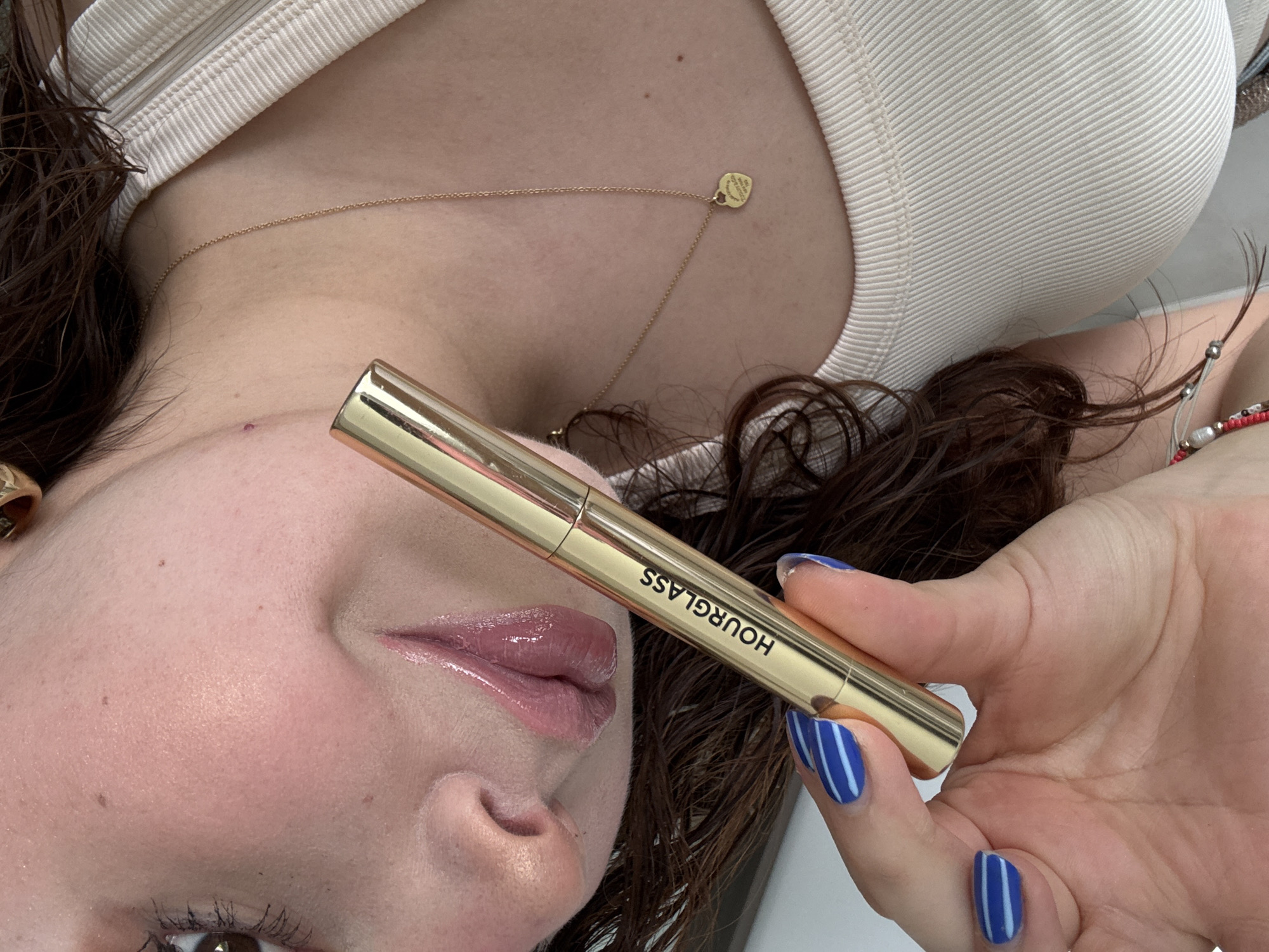 A selfie shows a woman holding a gold-colored tube labeled "HOURGLASS" near her face. Her skin appears to be fair, and she is wearing a shiny lip gloss. Her fingernails are painted blue with white stripes. She is wearing a light-colored top and a gold necklace. The background is slightly blurred, showing hints of her hair and what appears to be her surroundings. There is no information in the image that allows me to identify a location, city or country.