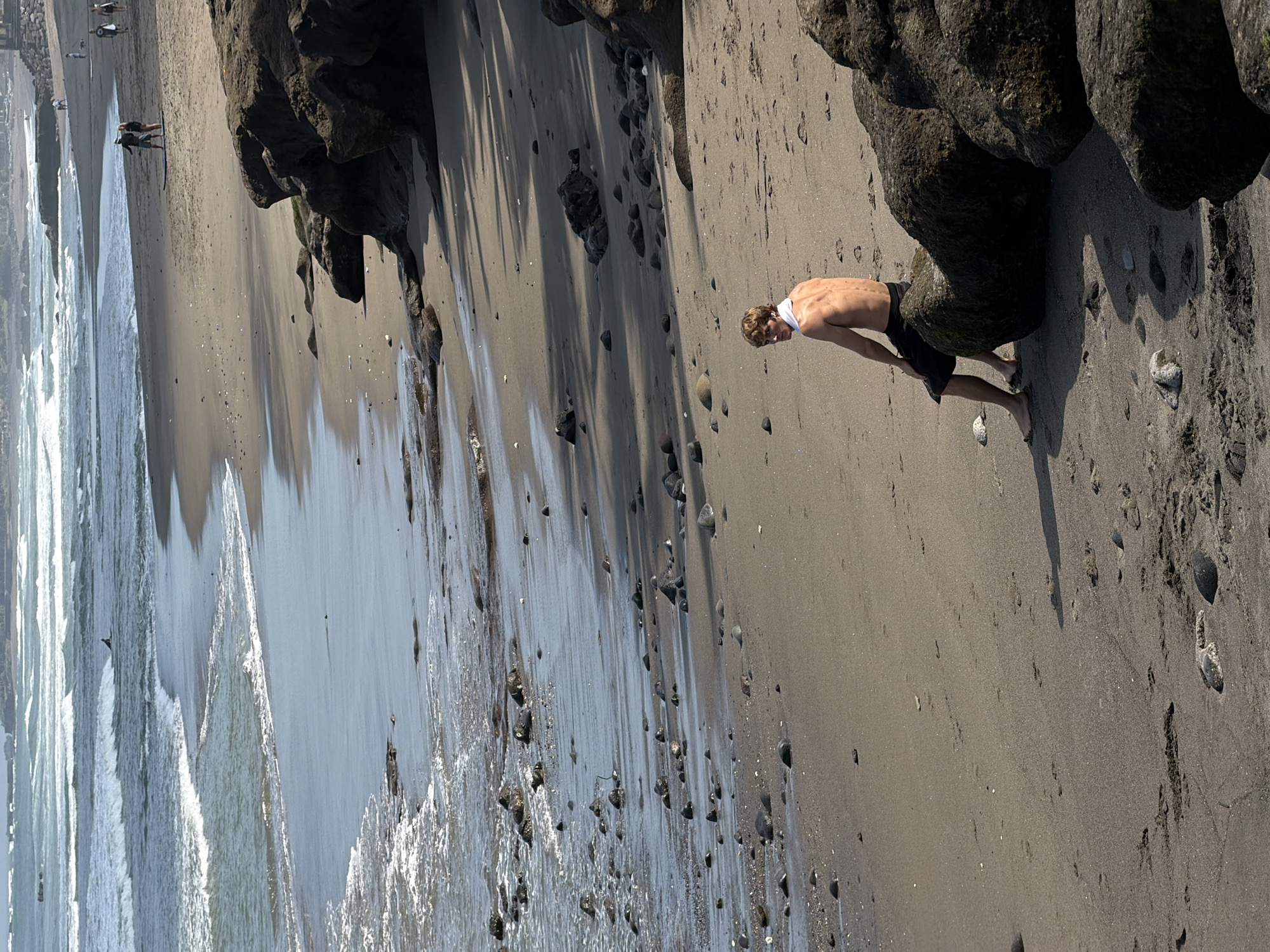 A young man with blond hair, wearing black shorts and a white towel around his neck, sits on a rock on a sandy beach. He is facing away from the camera, looking towards the ocean. The beach is strewn with rocks and pebbles, and the waves are crashing onto the shore. In the background, there are several other people walking on the beach and in the water. The sky is overcast, and the overall mood of the image is peaceful and serene. The location is North Kuta, Indonesia, a popular tourist destination known for its beautiful beaches and vibrant culture.