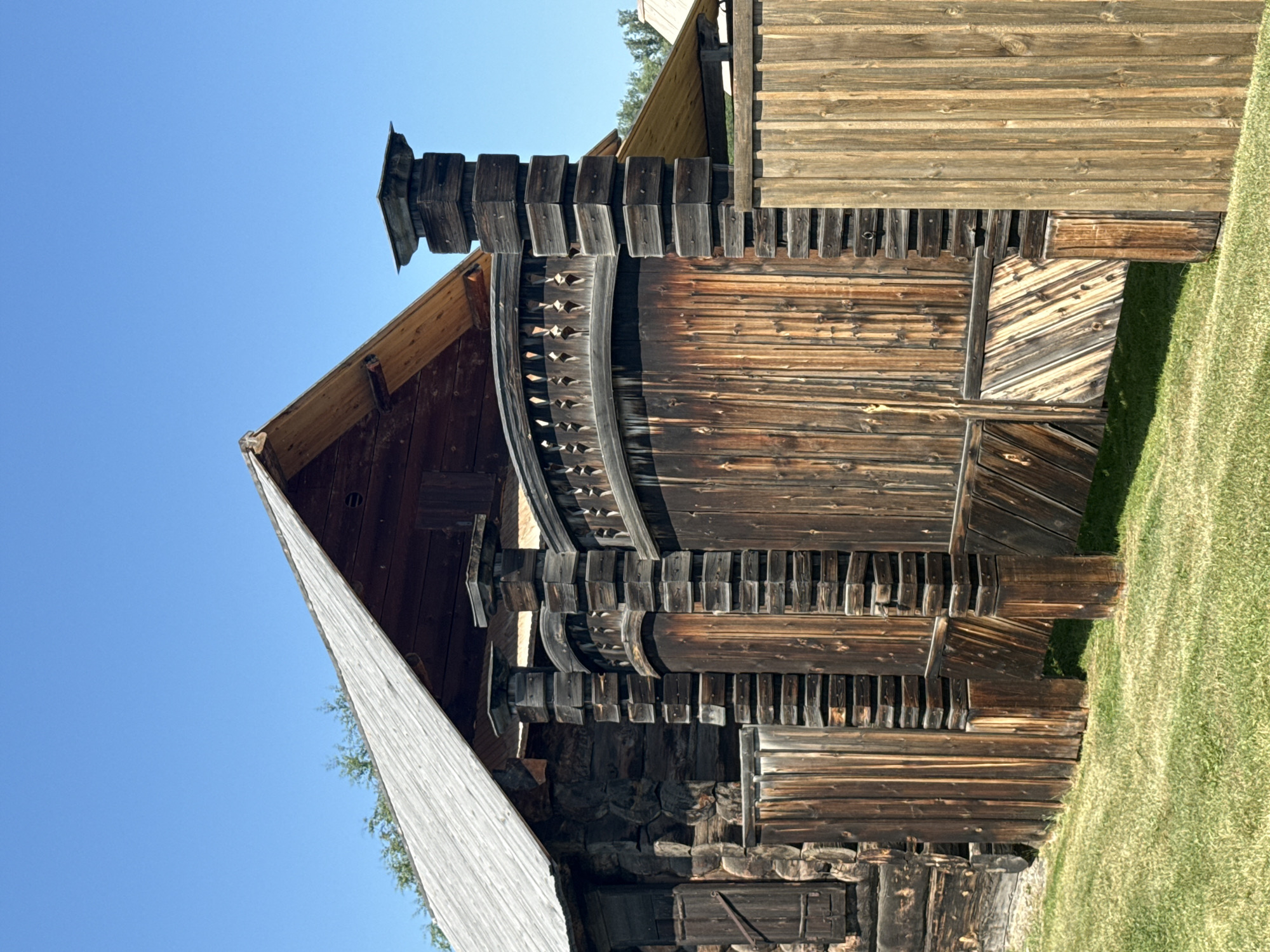 The image depicts a rustic wooden gate and part of a building under a clear blue sky in the city of Shushenskoye, Russia. The gate is elaborately constructed with vertical wooden planks, decorative carved elements along its top, and sturdy wooden posts. The building appears to be made of dark, aged logs, with a slanted wooden roof. The scene is set against a backdrop of green grass. There are no people or animals visible in the image. The overall impression is one of historical architecture in an outdoor setting.