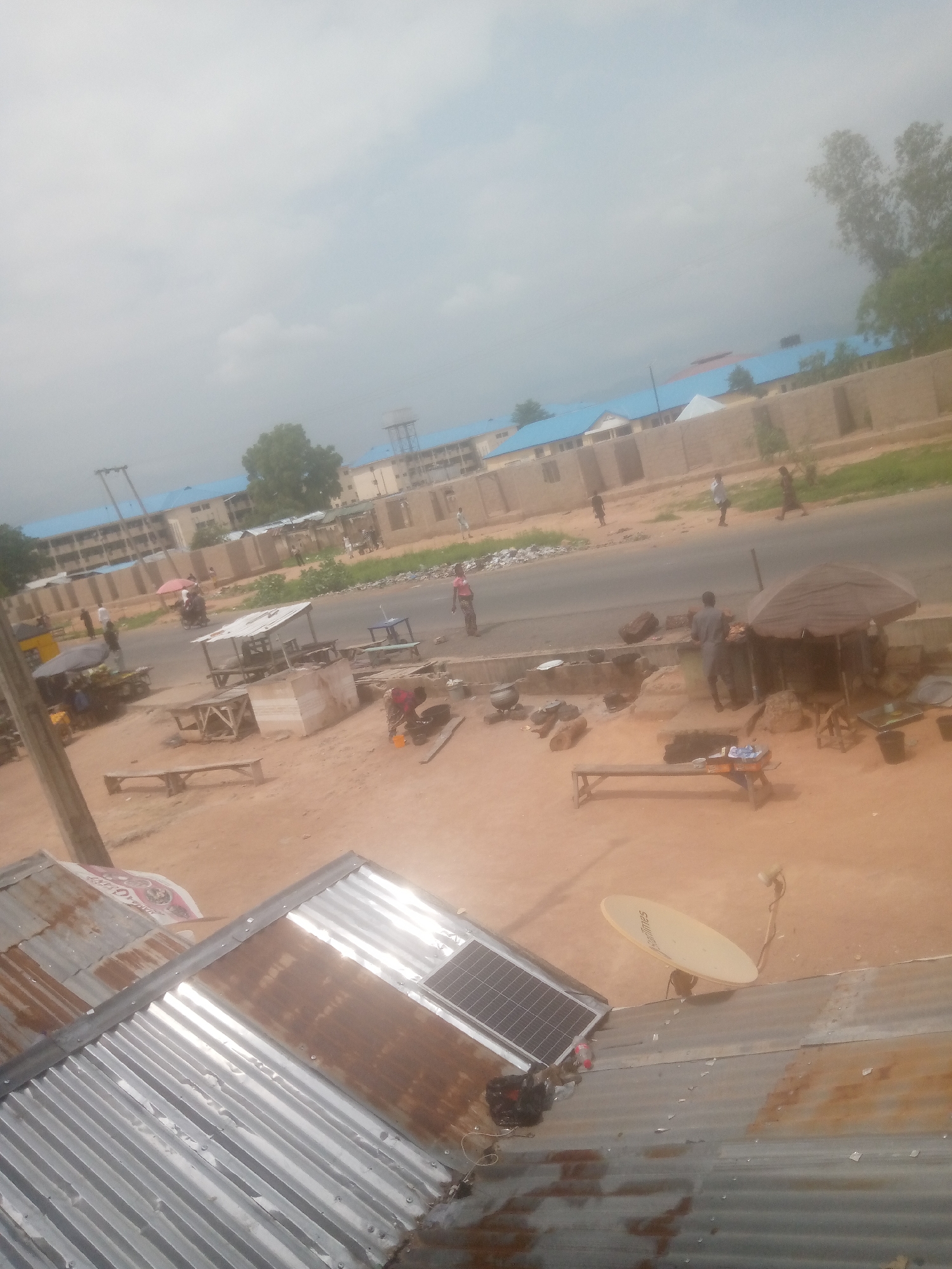 This image depicts a street scene in Jalingo, Nigeria, likely in an informal commercial or residential area. The foreground is dominated by corrugated metal roofs, some appearing weathered and rusty, suggesting a developing or established community. A solar panel is visible on one of the roofs, alongside a satellite dish.
The mid-ground shows a dusty, unpaved area bordering a paved road. Along the roadside, there are various informal stalls and structures. One stall has a thatched roof, providing shade over wooden tables and what appear to be cooking pots. Another stall, partially covered by a dark umbrella, features more cooking equipment and food preparation. Several people are present, engaged in activities such as cooking, standing, and walking. A person on a motorcycle with a pink umbrella is also visible. Benches are scattered throughout the area, and a few wooden tables are set up, indicating a place for commerce or rest.
In the background, a series of buildings with blue roofs are visible, possibly part of a school, residential complex, or public institution. A water tower stands behind these buildings. A low, light-colored brick wall runs along the front of these structures. People are seen walking on the road and sidewalk area. The overall environment is characterized by a blend of infrastructure, informal economic activity, and a relatively modest built environment, typical of many urban or semi-urban areas in Nigeria. The sky is overcast, suggesting potentially rainy or hazy weather.