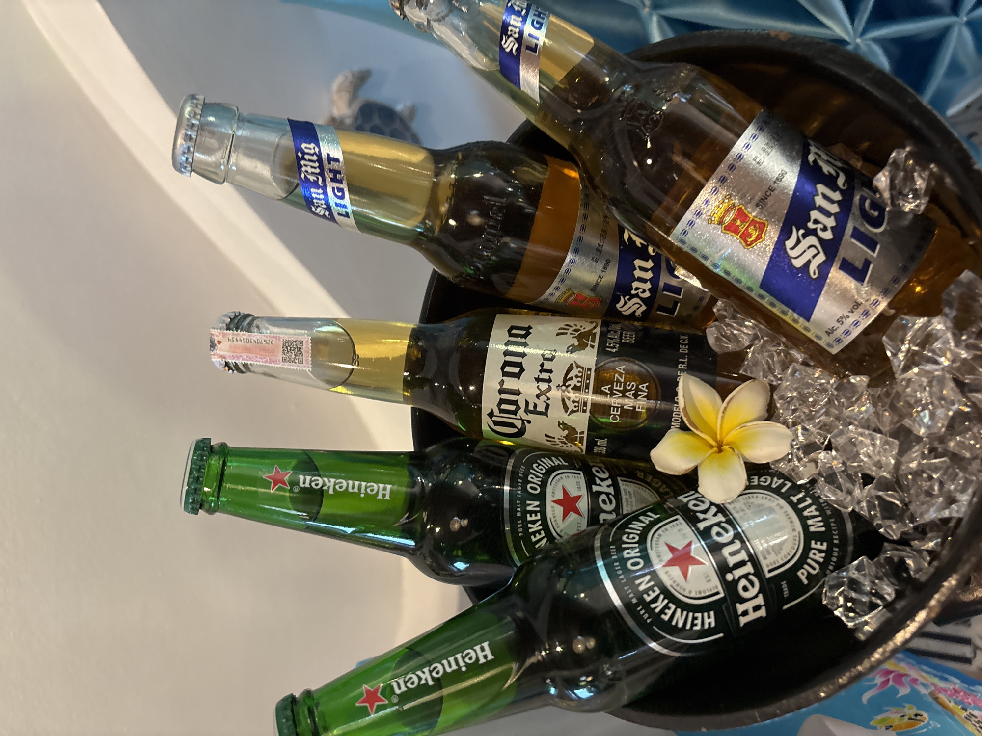 The image shows five bottles of beer in an ice-filled bucket. Three of the bottles are Heineken, one is Corona Extra, and two are San Miguel Light. A single yellow and white flower rests on top of the ice. The bucket is dark in color and appears to be made of metal or plastic. The background is a light-colored wall with some shadows. The overall scene suggests a casual gathering or celebration, possibly outdoors given the presence of ice and a flower. There are no people visible in the image. The location of Chalong, Thailand, is inferred from the presence of San Miguel Light, a popular beer in the Philippines, which is often consumed in tourist areas like Chalong, Thailand.