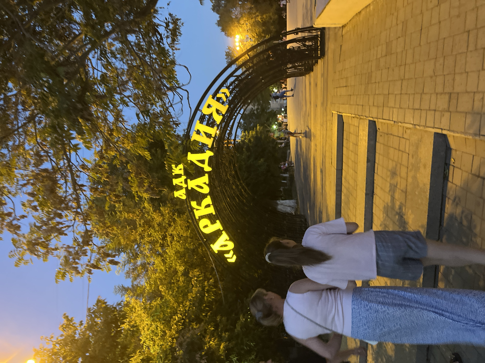 The image depicts a twilight scene at the entrance to Arkadiya Park in Astrakhan, Russia. A large, illuminated archway spells out "АРКАДИЯ" in bright yellow Cyrillic letters, welcoming visitors. The archway is constructed of dark metal and is adorned with foliage. In the foreground, two females, likely a mother and daughter, are walking away from the camera towards the park. The adult female is wearing a white sleeveless top and a long blue patterned skirt, carrying a white crossbody bag. The younger female is wearing a pink t-shirt and grey shorts. The pathway leading into the park is paved with light-colored bricks, and illuminated by warm streetlights casting long shadows. The park itself is lush with trees, their leaves glowing in the artificial light. Other park-goers are visible in the background, suggesting a pleasant evening atmosphere. The sky is a deep blue, indicating the transition from day to night.