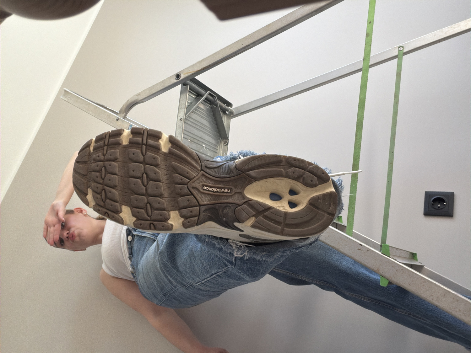 An extreme low-angle indoor shot captures a person seen from directly below, positioned above the camera. The sole of a brown and beige New Balance sneaker dominates the foreground, with the brand name "new balance" visible on its traction pattern. The person, wearing a white t-shirt and ripped blue denim jeans, is visible looking down towards the camera with a playful, pouting expression, partially obscuring their face with their hand.
Behind the person, a silver-colored metal folding ladder is prominent, suggesting the individual might be on or leaning against it. The background features a plain, light-colored wall, possibly white or light grey. On the right side of the frame, a black electrical outlet with two circular holes is mounted on the wall. A thin green strap or tape hangs vertically in the mid-ground, near the ladder. The image has a lighthearted and candid feel, with the unique perspective placing the viewer directly beneath the subject. The setting is an indoor space, consistent with a location in Dreiliņi, Latvia.