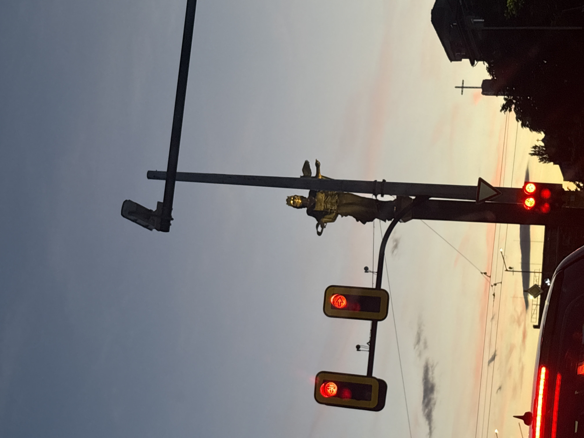 In an urban environment during twilight, a statue of a golden figure wearing a crown stands prominently on a lamppost. Two traffic lights with red signals are visible to the left of the statue. Further to the right, a building with a cross on top can be seen silhouetted against the colorful sky. The foreground is partially obscured by the top of a vehicle, its brake lights illuminated in red. The scene is characterized by a blend of man-made structures and natural light from the setting sun.