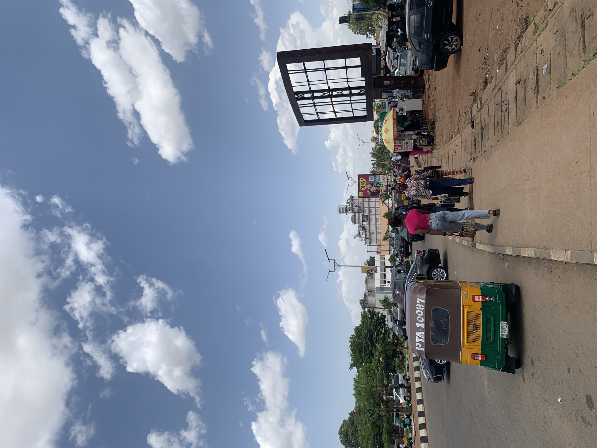 The image depicts a vibrant street scene on a sunny day in Jos, Nigeria, characterized by urban activity and clear skies. The perspective is from the side of a road, looking down a street lined with commercial establishments and pedestrians.
In the foreground, a green and yellow tricycle, commonly known as a "Keke Napep," is visible on the paved road, with "PTA-10087" and license plate "RSH-478YQ" clearly marked on its rear. To its left, a concrete curb separates the road from a pedestrian walkway. Several individuals are walking along this walkway, some carrying bags, indicating daily errands or commutes.
The midground features more people, some clustered around street vendors. A prominent yellow umbrella, likely belonging to a stall, displays the text "POS" (Point of Sale) and "MAGGI." Another sign nearby shows the "MTN" logo, a telecommunications company. Behind these activities, a large, rectangular metal structure resembling an empty billboard frame stands tall against the sky. Several cars are parked or moving along the street further down.
The background shows various urban buildings, some multi-story, interspersed with green trees. The sky dominates a significant portion of the image, appearing bright blue with scattered, fluffy white cumulus clouds, suggesting fair weather and daytime. The overall impression is one of a bustling city street on a clear day, filled with everyday life and commercial activity.