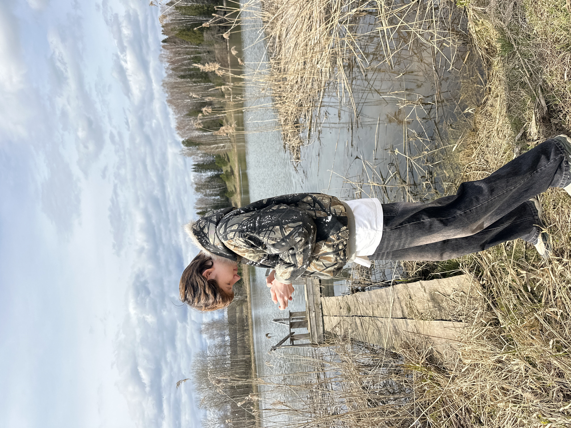 The image shows a young person standing on a wooden dock beside a calm body of water, likely a lake or pond, in what appears to be a rural setting in Альняш, Russia. The individual, who appears male with short brown hair, is seen in profile, looking down towards the water with their hands clasped together in front. They are wearing a camouflage-patterned jacket with a light-colored, possibly fur-lined, trim around the hood or collar, a white undershirt, and dark jeans.
The scene is characterized by natural elements. The water's edge and the areas around the wooden dock are lined with abundant dry reeds and tall grasses, suggesting late autumn or early spring. Across the water, a dense forest line composed primarily of bare deciduous trees with some scattered evergreens stretches towards the horizon. A small, simple wooden structure, possibly a part of the dock system or a standalone platform, is visible further out on the water.
The sky is cloudy and light grey, indicating daytime with diffused natural light, though it doesn't appear heavily overcast. The overall mood is tranquil and contemplative, with the person seemingly observing the water or their immediate surroundings. No specific activity like fishing is depicted. There is no visible text in the image.
