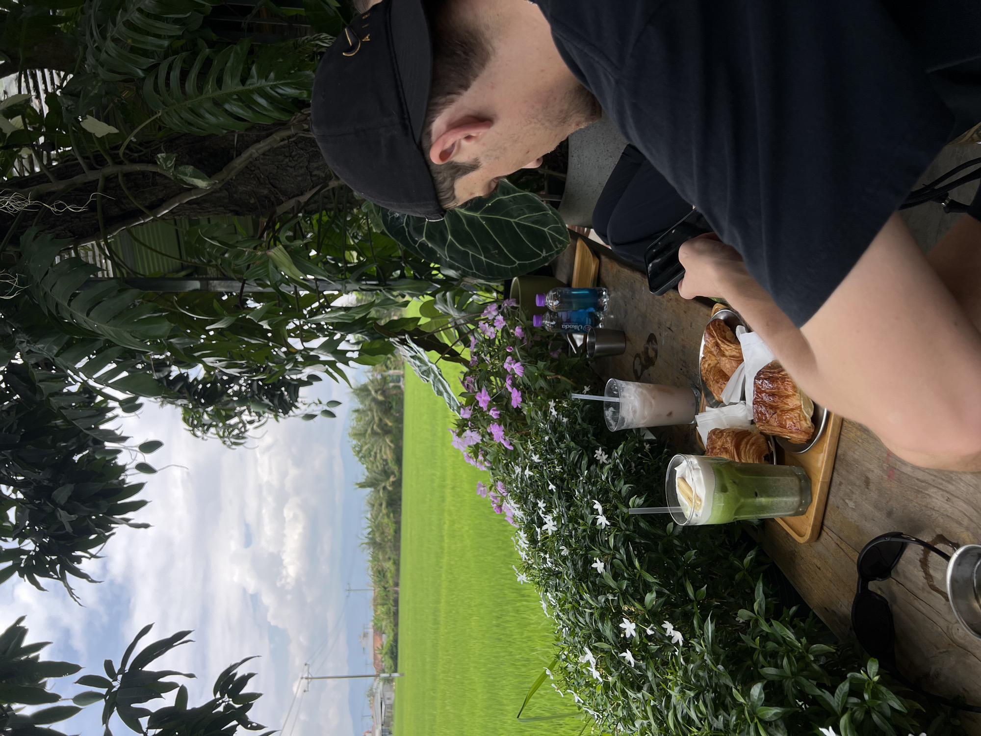 A man in a black shirt and cap is seated at a wooden table with a view overlooking a rice field. The table is set with croissants on a tray, two drinks, small bottles of Okado mineral water, and sunglasses. The scene is framed by foliage with white and purple flowers. The location is implied to be in Diên Khánh, Vietnam, based on the rice paddy landscape. The man is looking down at his phone, which he is holding in his hand.