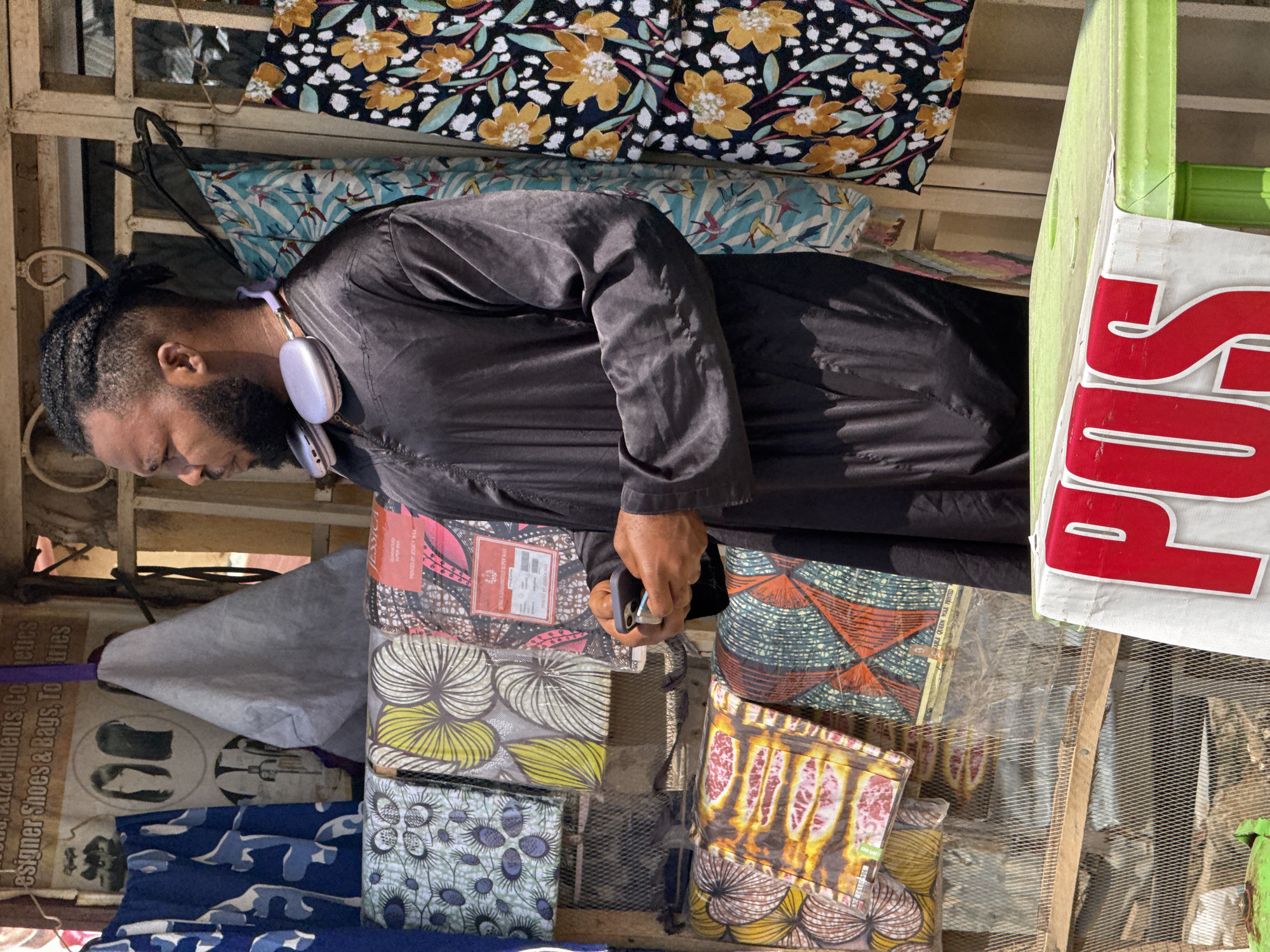 The image shows a man standing in front of a stall selling fabrics, likely in Abuja, Nigeria. The man is wearing a black traditional garment and has headphones around his neck. He is holding a mobile phone in his hands and appears to be looking at it. The stall displays a variety of colorful fabrics with different patterns. In the foreground, a green table is visible with a white cloth on it, bearing the red letters "POS". The background shows a weathered structure, possibly part of a market or street vendor setup. The lighting suggests it is daytime.