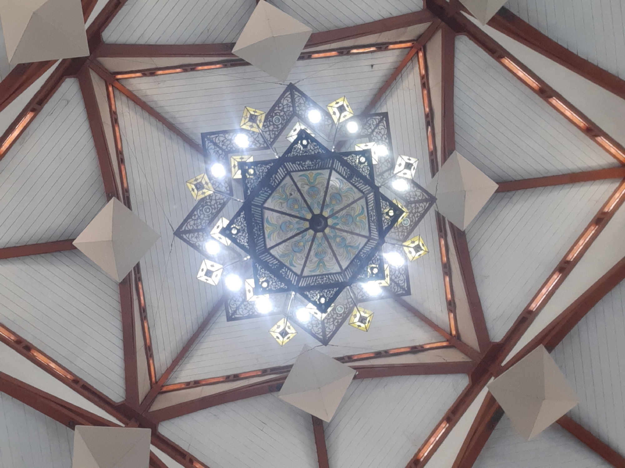 The image shows a low-angle view of a geometrically intricate chandelier hanging from a wooden and white-paneled ceiling. The chandelier is a complex, multi-tiered structure with a central octagonal section featuring decorative metalwork and colorful glass panels. Around this central piece, smaller, square-shaped lamps with yellow glass are arranged in a starburst pattern, interspersed with what appear to be small, angular light fixtures. The ceiling itself is constructed of wooden beams radiating outwards from a central point, supporting white wooden panels. Several three-dimensional, pyramidal decorative elements are attached to the ceiling, positioned between the beams. The lighting is bright, originating from the chandelier, which illuminates the surrounding ceiling structure. The image appears to be taken indoors, possibly in a public building such as a mosque or a cultural center, given the ornate design of the chandelier and the architectural style of the ceiling. The location is identified as a mosque in Bandung City, Indonesia.