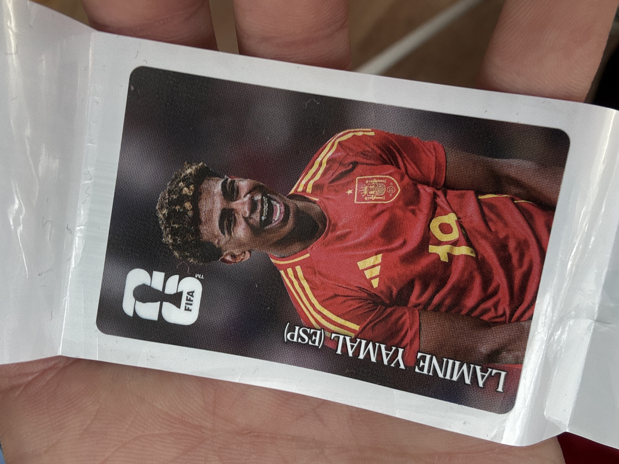 An image shows a trading card with a portrait of a young man wearing a red soccer jersey with yellow stripes. The jersey has a crest on the chest and the number 10 on the front. The man has curly hair and is smiling, showing braces on his teeth. The card also features the FIFA logo and the text "LAMINE YAMAL (ESP)". The card is held by a person, with parts of their fingers visible on the left and right sides of the image. The background of the card is dark. The scene appears to be indoors, with lighting that highlights the card.