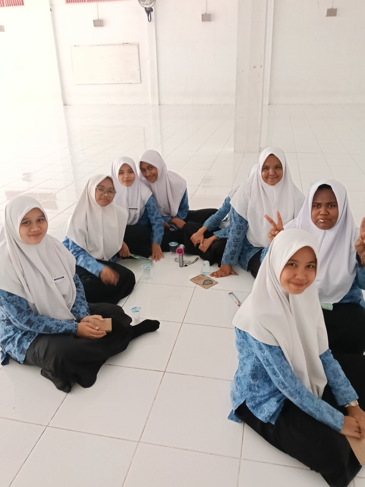A group of seven young women wearing white headscarves and blue patterned shirts are sitting on a white tiled floor indoors. The room appears to be in a public building, possibly a school, located in Lhokseumawe, Indonesia. The women are arranged in a semi-circle, and there are a few small objects, including a glass and what appears to be a small electronic device, placed on the floor in front of them. Two of the women are making the peace sign with their fingers.