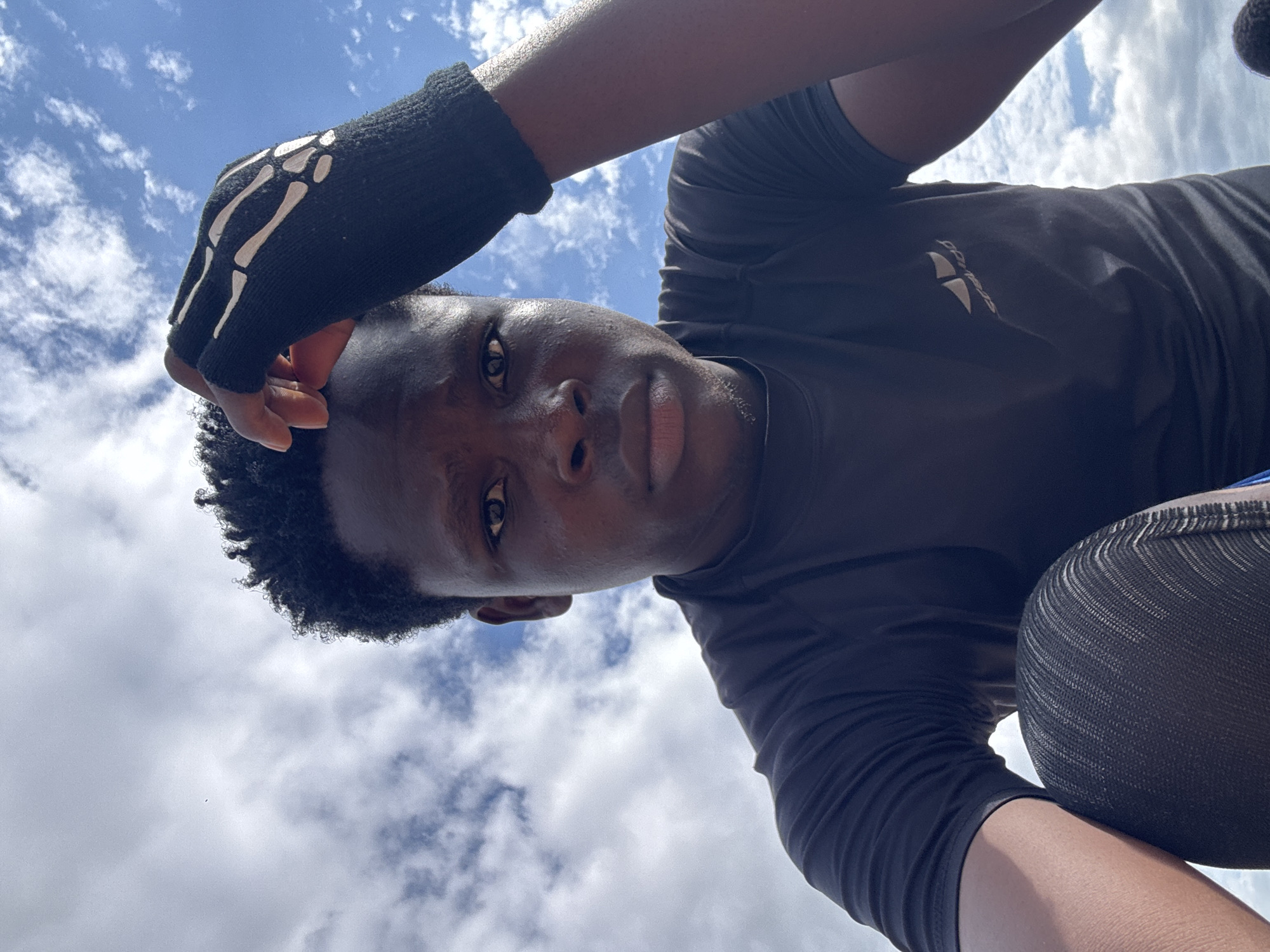The media file is a selfie taken from a low angle, looking up at the sky. The subject is a young Black person with curly hair, wearing a black t-shirt and a black fingerless glove with a white skeleton hand design. They are also wearing athletic leggings. The background is a bright blue sky with scattered white clouds, suggesting daytime with fair weather. The person is looking directly at the camera, with a neutral expression. The image appears to be taken outdoors in Ihiagwa, Nigeria. The clothing suggests the person might be engaged in physical activity or sports. There is a visible logo on the t-shirt, but the text is not legible.