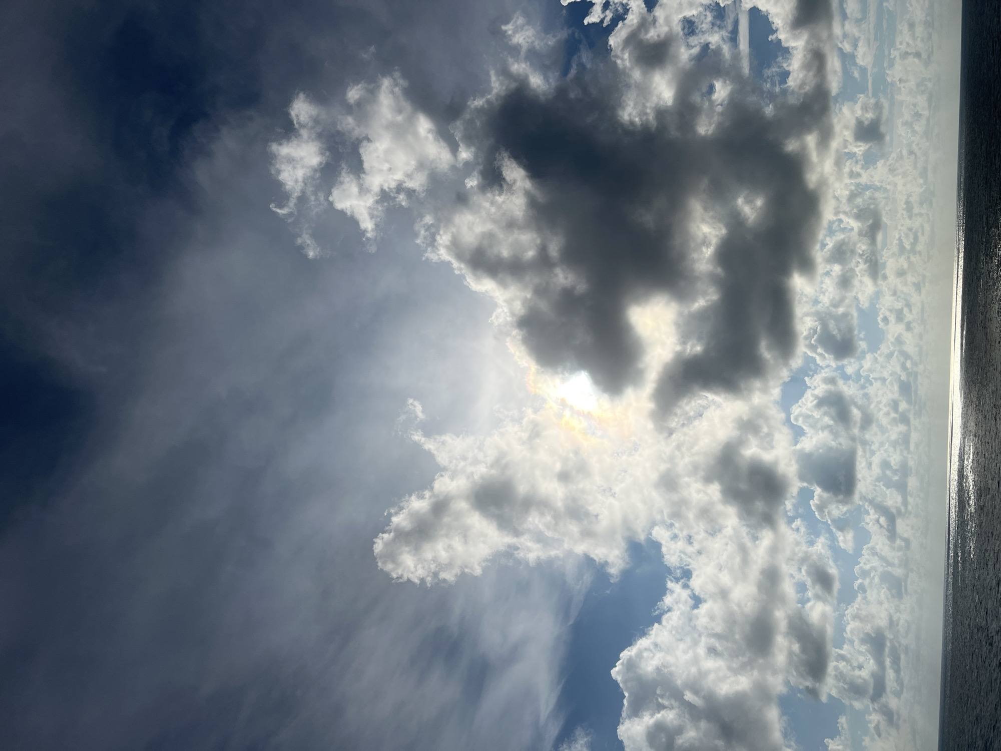 A wide shot captures a vast expanse of the ocean under a dramatic sky filled with a mix of dark and light clouds. The sun peeks through a gap in the clouds, casting a bright, diffused light that creates a shimmering effect on the dark, rippling water. The horizon line is sharp, dividing the deep blue-grey sea from the dynamic sky. The clouds are varied in texture and density, with some appearing wispy and thin, while others are thick and opaque, casting shadows that add depth and contrast to the scene. The overall impression is one of natural beauty and the power of the elements.