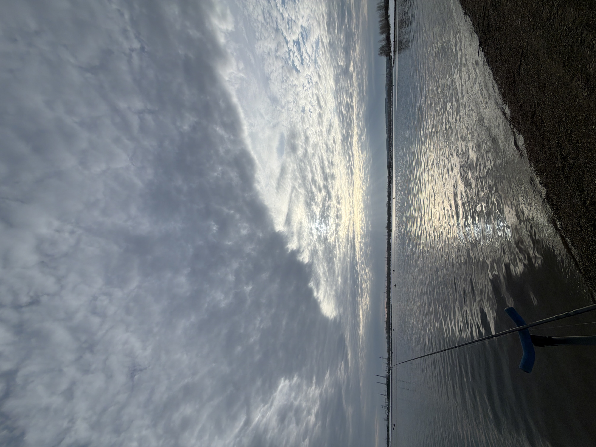 The image depicts a scene from Chaikovsky, Russia. The view is oriented towards the horizon where the sky meets a body of water, likely a river or large lake. The sky is filled with a mix of dark, dense clouds and brighter, thin cloud formations reflecting sunlight. The water surface shows ripples and reflections of the sky, with the sun creating shimmering patterns. A shoreline is visible on the right side, composed of dark gravel or similar material. A fishing rod extends into the water from the right foreground, indicating someone fishing. Power lines are visible in the distance, suggesting an urbanized area. The overall lighting suggests either early morning or late afternoon.