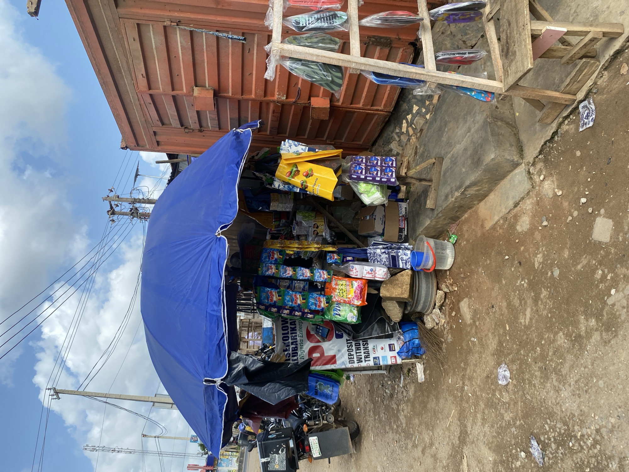 A street-side kiosk in Lagos, Nigeria, is sheltered by a blue umbrella. The kiosk is constructed of wooden shelving, overflowing with various consumer goods, from packaged foods to household items, alongside advertisements for financial services. A parked motorcycle sits to the side of the kiosk. The ground around the kiosk is dirt. A rusty metal container serves as a backdrop, with utility poles and wires visible in the sky above.