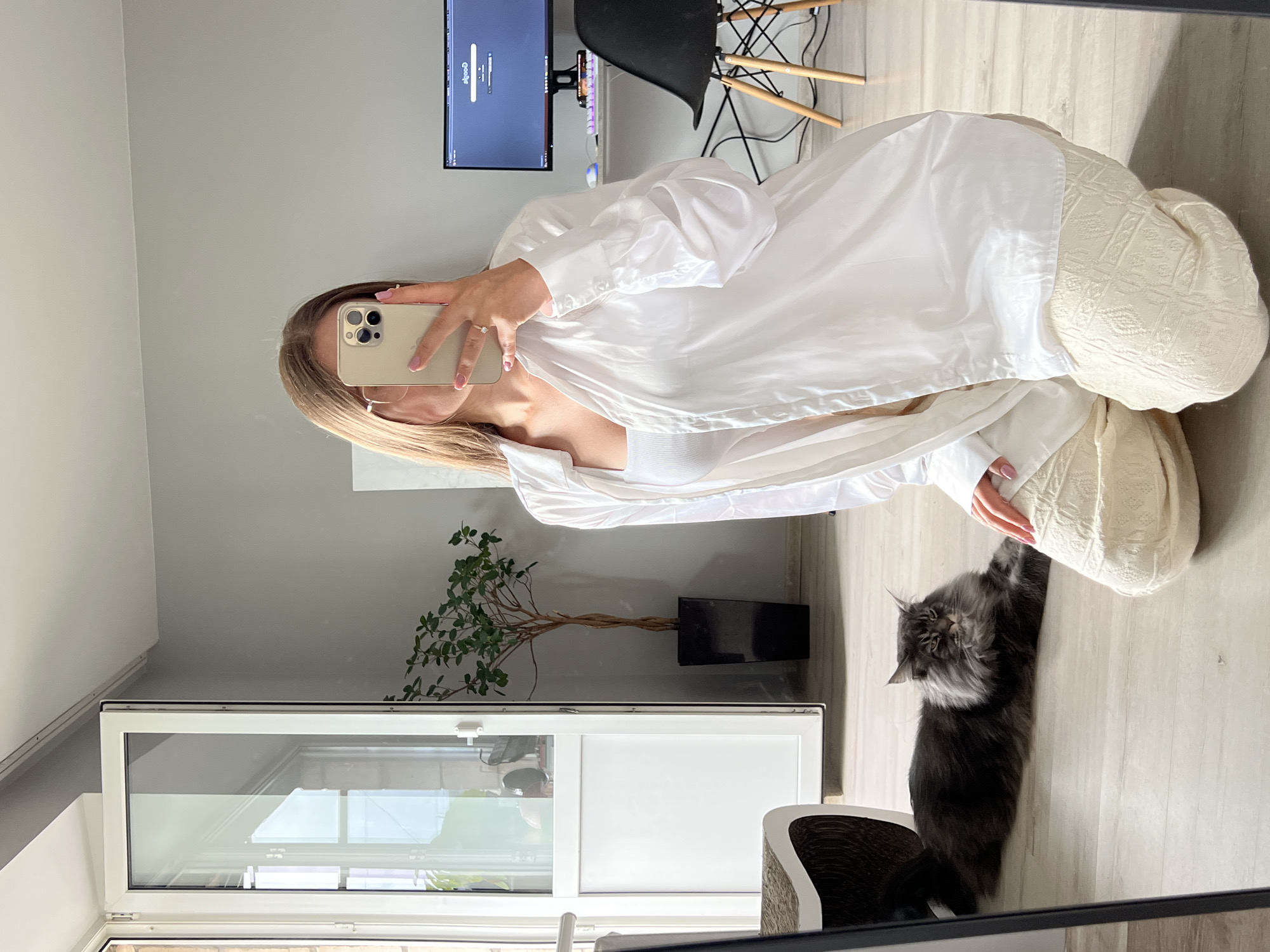 A young woman with long blonde hair is taking a mirror selfie while kneeling on a wooden floor. She is wearing a white camisole and a white oversized button-up shirt, and cream-colored patterned pants. In the reflection, a large grey Maine Coon cat is lying on the floor next to her. Behind the woman, a modern room is visible with a desk, a computer monitor, a black chair, and a tall potted plant in a black rectangular planter. To the left, a large white door leading outside is open, revealing part of a balcony and city views. The scene appears to be an apartment in Ternopil, Ukraine, likely in a modern building, possibly with a balcony overlooking the city.
