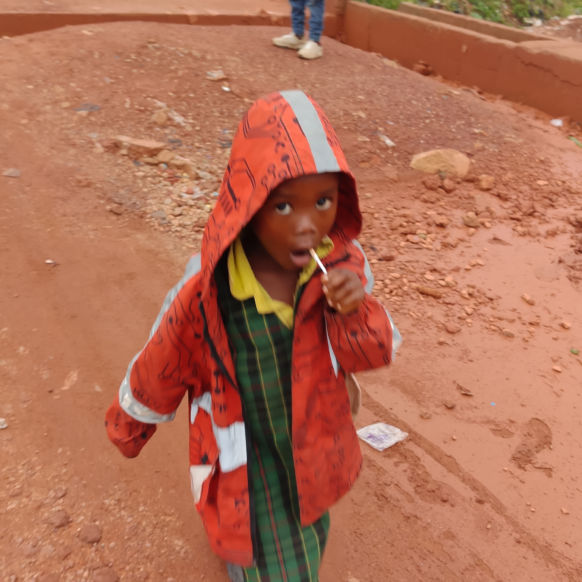 A young child, appearing to be a boy, stands on a dirt road with reddish-brown soil and small stones. He is wearing a bright orange hooded jacket with a reflective stripe on the hood and black patterns, over a green and black plaid shirt and a yellow collar. The child is holding a white lollipop to his mouth. In the background, a person wearing blue jeans and white shoes is partially visible standing on an elevated dirt embankment. The environment suggests an outdoor, possibly rural or semi-urban setting in Gura Top, Nigeria, characterized by the unpaved ground and the presence of rudimentary infrastructure. A crumpled white plastic bag is on the ground to the child's right.