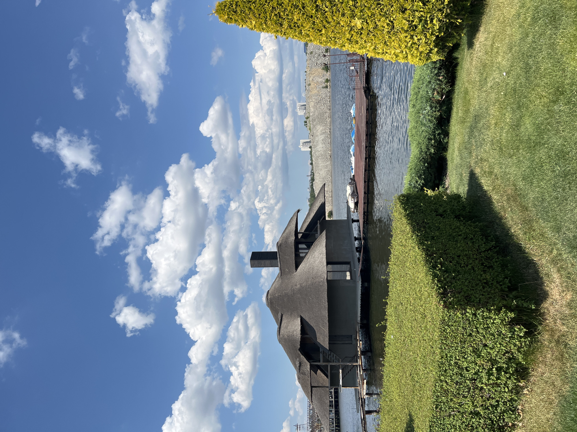 This image depicts a scenic view of a modern, thatched-roof building situated on stilts over a body of water. The building features a dark grey exterior and a complex, multi-peaked roof, with a prominent black chimney. Wooden walkways and piers surround the structure, with several small paddle boats docked on one of the piers. In the foreground, meticulously manicured green bushes and a well-maintained lawn are visible.
The background reveals a rugged, grey stone embankment rising from the water, with several modern white buildings situated on its crest. A bright yellow evergreen tree stands tall on the right side of the frame, adding a vibrant contrast to the scene. The sky is a clear blue, dotted with fluffy white clouds, suggesting a sunny day.
This location is identified as the "Bar" district of the restaurant complex "Big Five" in Dnipro, Ukraine. The restaurant complex is situated on the Dnipro River, near the former river port.
