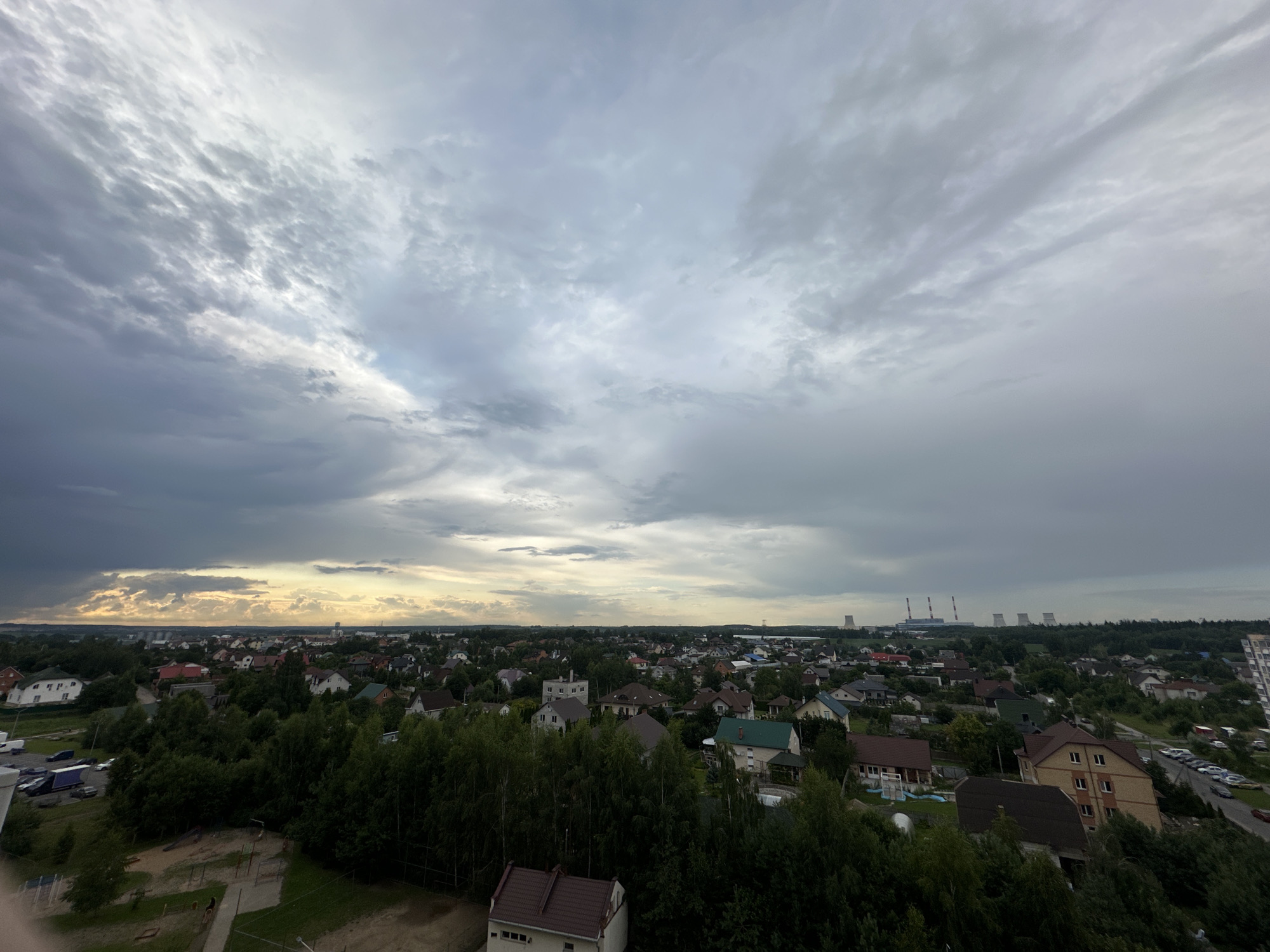 An aerial, wide-angle view captures a suburban landscape under a dramatic, cloudy sky. The foreground is dominated by the dark green canopy of dense trees, with a playground visible in the lower left quadrant. Beyond the trees, a collection of residential houses with varied roof colors and architectural styles stretches across the middle ground. Some houses have light-colored facades, while others are darker. Roads weave through the neighborhood, with several parked cars visible along one. In the distance, industrial structures, including cooling towers of a power plant with tall smokestacks, are silhouetted against the horizon. The sky is a dynamic mix of dark gray storm clouds on the left, transitioning to lighter, scattered clouds with patches of yellow and white light suggesting a break in the clouds or sunset on the right. The overall scene depicts a residential area on the outskirts of a city, with evidence of industrial activity in the background.