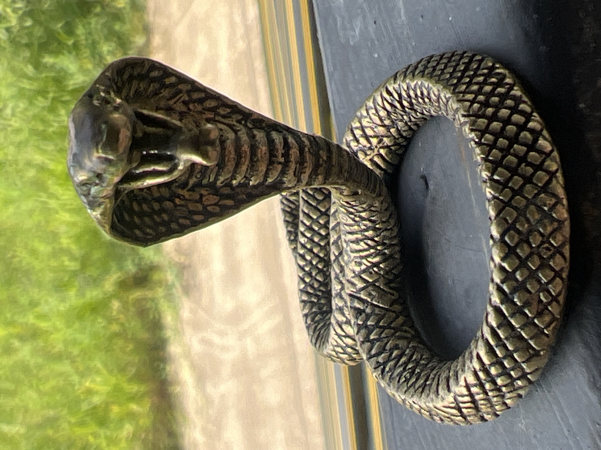 A bronze figurine of a cobra is coiled on a dark, flat surface. The cobra's head is raised, its hood flared, and its mouth open. The figurine is detailed, with textured scales. In the blurred background, green foliage is visible above a sandy or gravelly path, suggesting an outdoor setting, potentially near vegetation. The figurine is positioned near a window or edge, as indicated by horizontal lines in the background that could be window panes or the edge of a ledge. The image's lighting suggests it was taken during the day. There is no indication of Karon, Thailand, in the image itself.