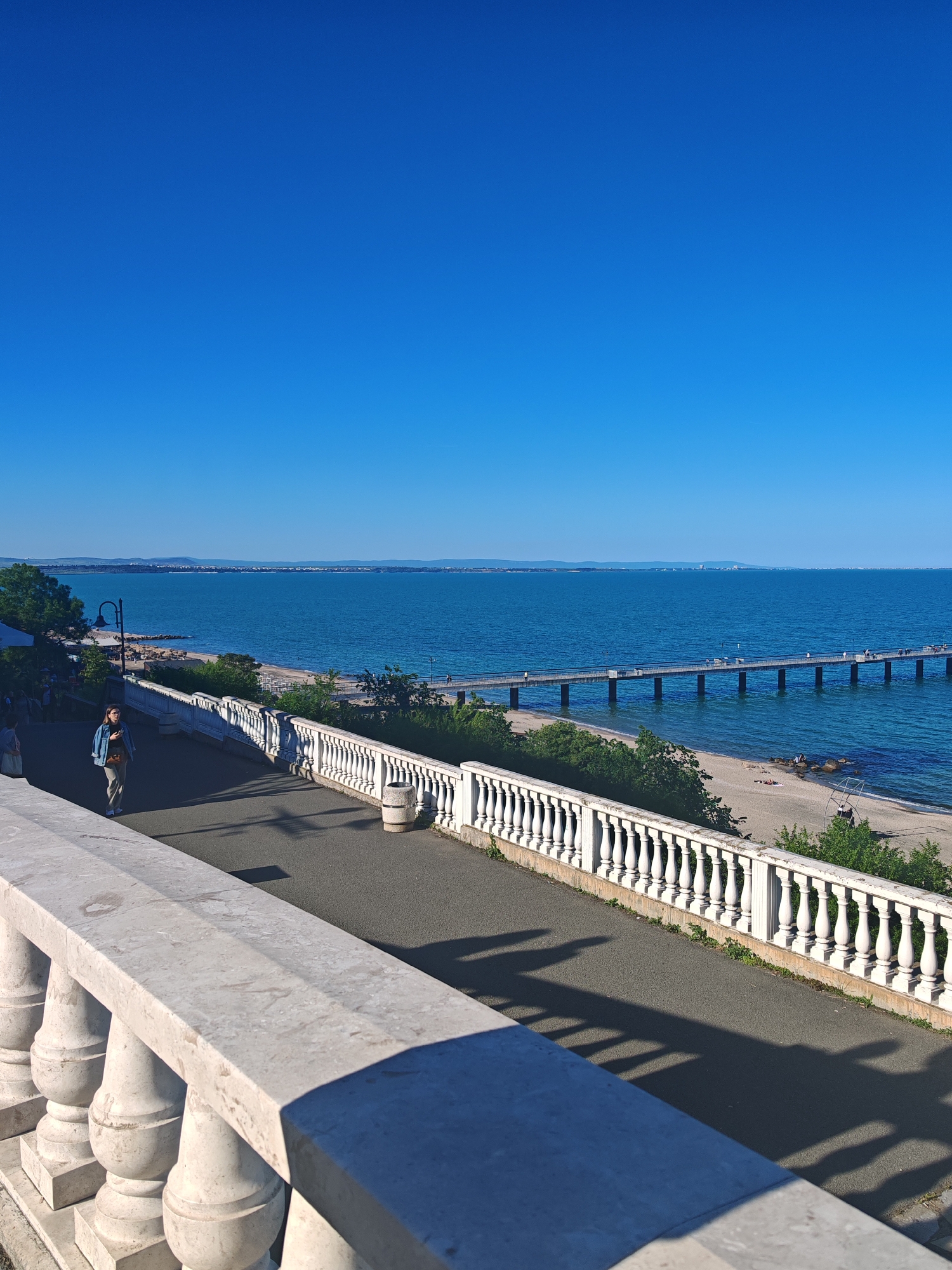 The image depicts a sunny day in Burgas, Bulgaria, with a view of the Black Sea. A long pier extends into the water, dotted with people. A walkway with a white balustrade runs parallel to the shore. A woman walks along the walkway. Below the walkway is a beach with some people. The sky is a clear, deep blue.