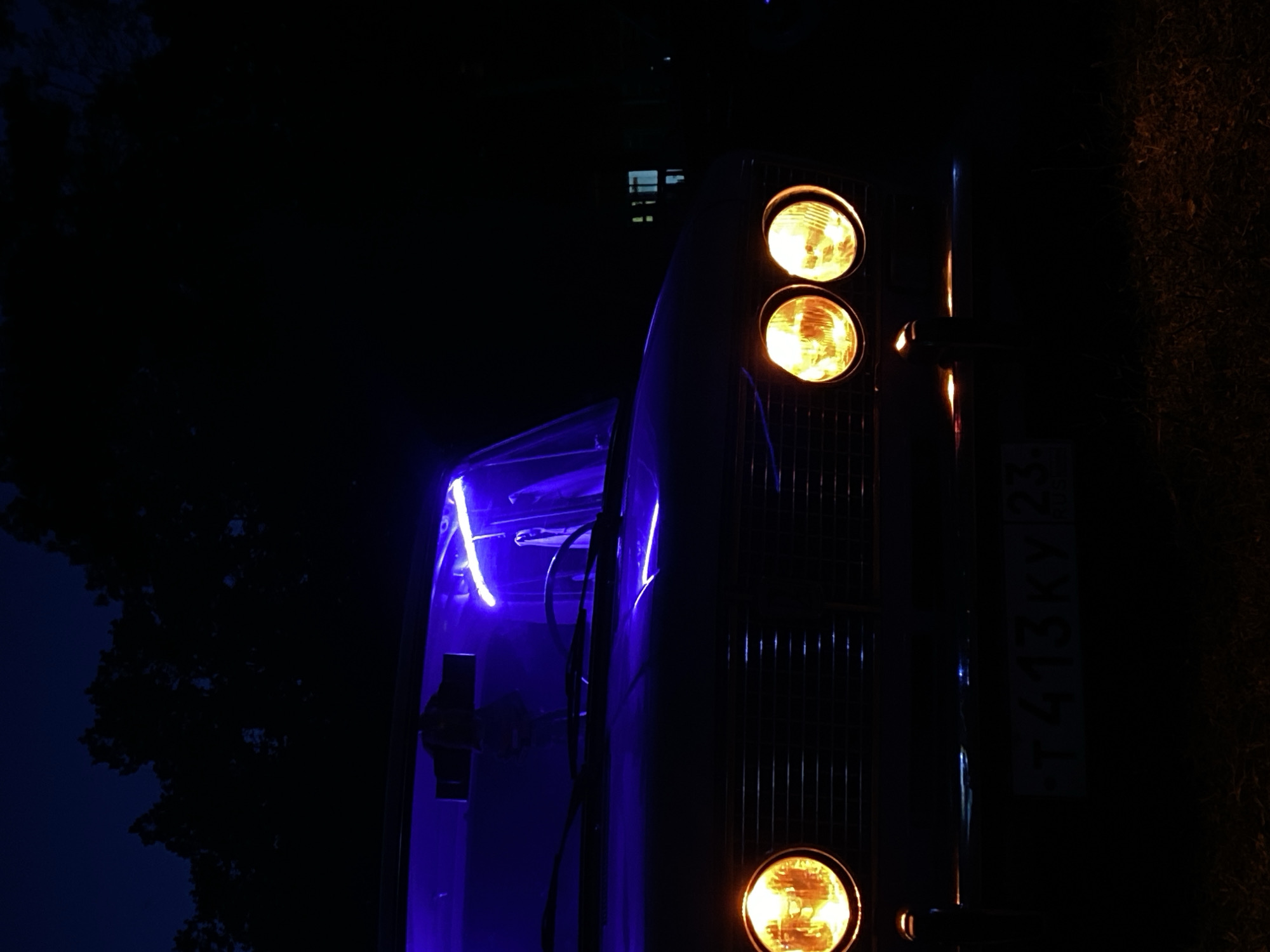 A vintage car is parked at night in Bezymyannoye, Russia. The car's headlights are illuminated with a warm yellow glow, and a neon blue light is visible inside the windshield. The car's grille is dark, and the license plate is partially obscured. In the background, silhouetted trees are visible against a dark night sky. A building with illuminated windows can be seen in the far distance. The ground is covered in grass and fallen leaves. The scene is dimly lit, with the car's lights providing the primary source of illumination.