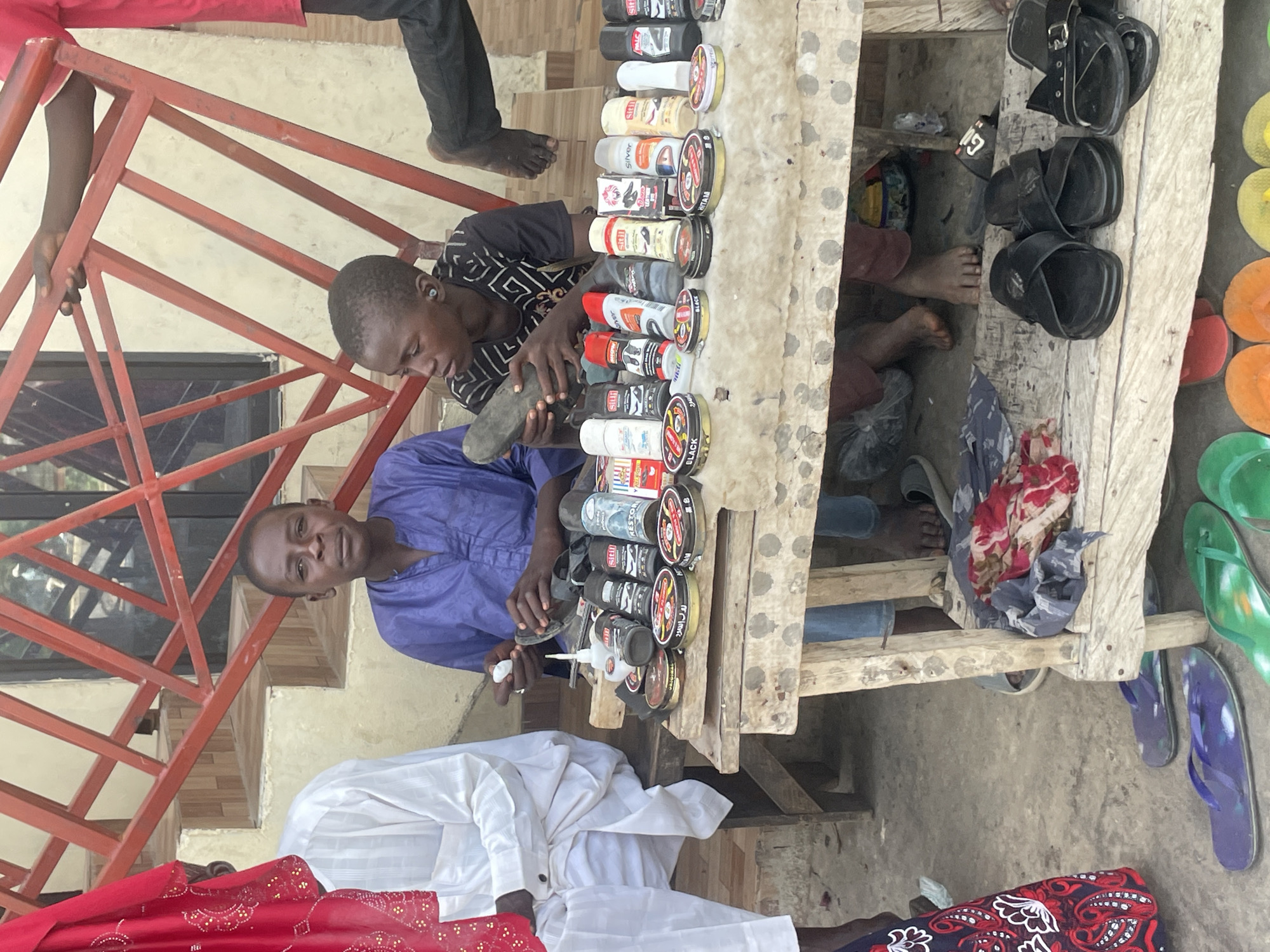 The image depicts a street scene in Bauchi, Nigeria, where a shoe-shining business is in operation. Two young men are the primary subjects. One, wearing a black and white patterned t-shirt, is actively shining a shoe. He is leaning over a wooden stall laden with various shoe polish tins, sprays, and brushes. The other young man, dressed in a vibrant purple traditional outfit, is seated and looking towards the viewer. In the foreground, a person wearing a white traditional garment is also visible, along with a portion of a red and white patterned fabric.
The stall itself is a makeshift wooden structure, suggesting an informal marketplace or roadside vendor. Various black sandals are displayed on the stall and to its right, some on a wooden plank. Several pairs of colorful flip-flops, including green and purple, are visible on the ground near the stall. The setting appears to be outdoors, likely during daylight hours, though the exact time of day and weather conditions are not explicitly discernible. The overall atmosphere suggests a busy, active commercial exchange common in many Nigerian urban areas. The young men appear focused on their tasks, with the one shining the shoe showing concentration. The scene conveys a sense of everyday commerce and livelihood.