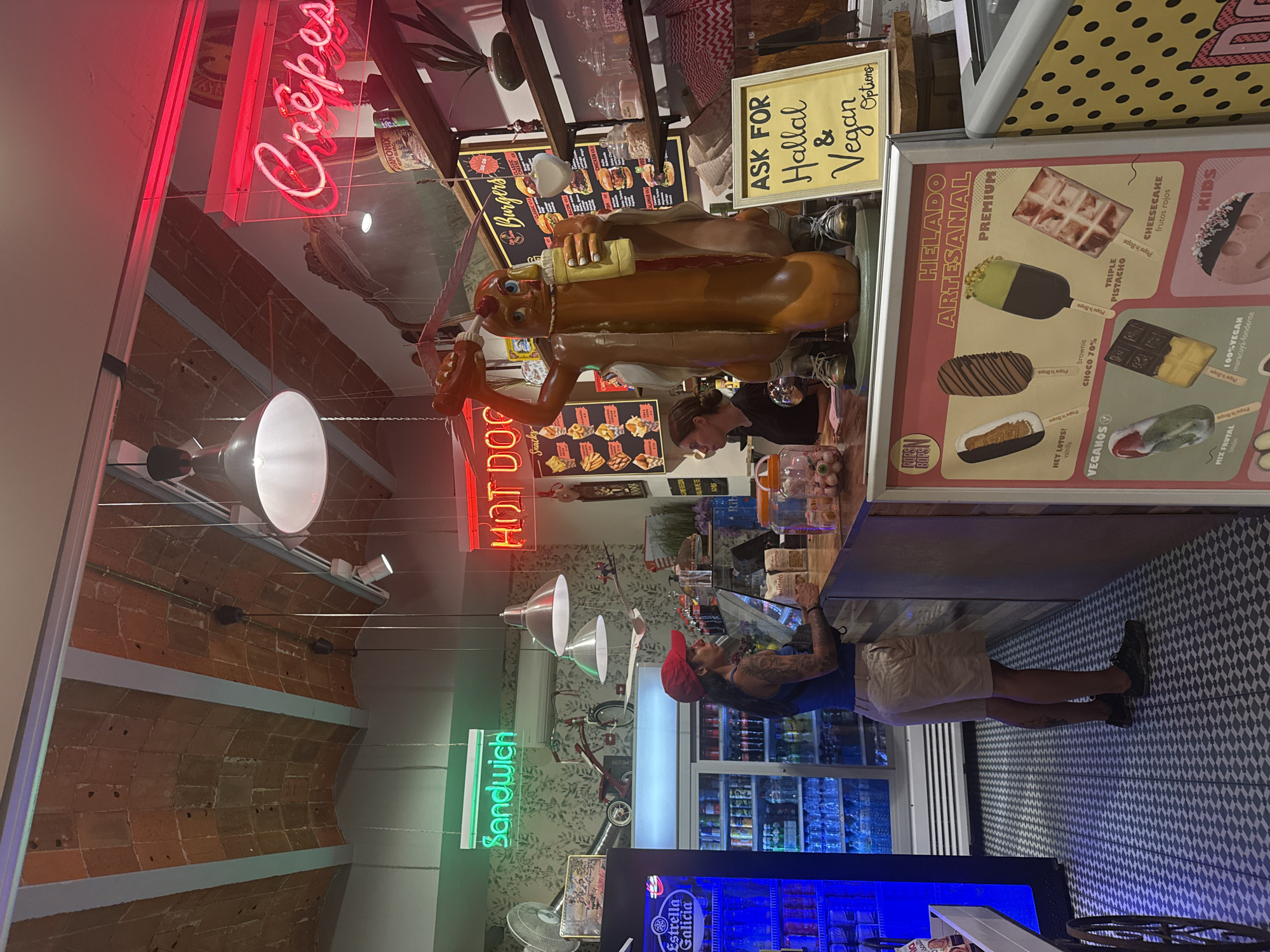 The image shows the interior of a casual food establishment in Tossa de Mar, Spain, likely a snack bar or ice cream shop. The establishment features a rustic ceiling with exposed brickwork and modern industrial lighting. In the foreground, a large, anthropomorphic hot dog statue is prominently displayed, holding a hot dog and a bottle of mustard. A sign next to it reads "ASK FOR Hallal & Vegan Options."
A woman with tattoos on her arms and wearing a red cap and khaki shorts stands behind a wooden counter, interacting with a customer who is partially visible on the left. Behind her, a refrigerated display case is filled with beverages, and a neon sign above it reads "Sandwich." To the right, another neon sign glows red, indicating "HOT DOGS." Further to the right, shelves are stocked with various items, including jars of candy and food packaging. A framed menu board displays options for "Burgers."
In the lower right quadrant, a framed advertisement for "HELADO ARTESANAL" (artisanal ice cream) is visible, showcasing different ice cream flavors and types, including "VEGANOS" (vegan options) and "KIDS." The floor is covered in a patterned tile. The overall atmosphere is bright and informal, characteristic of a tourist-oriented eatery.